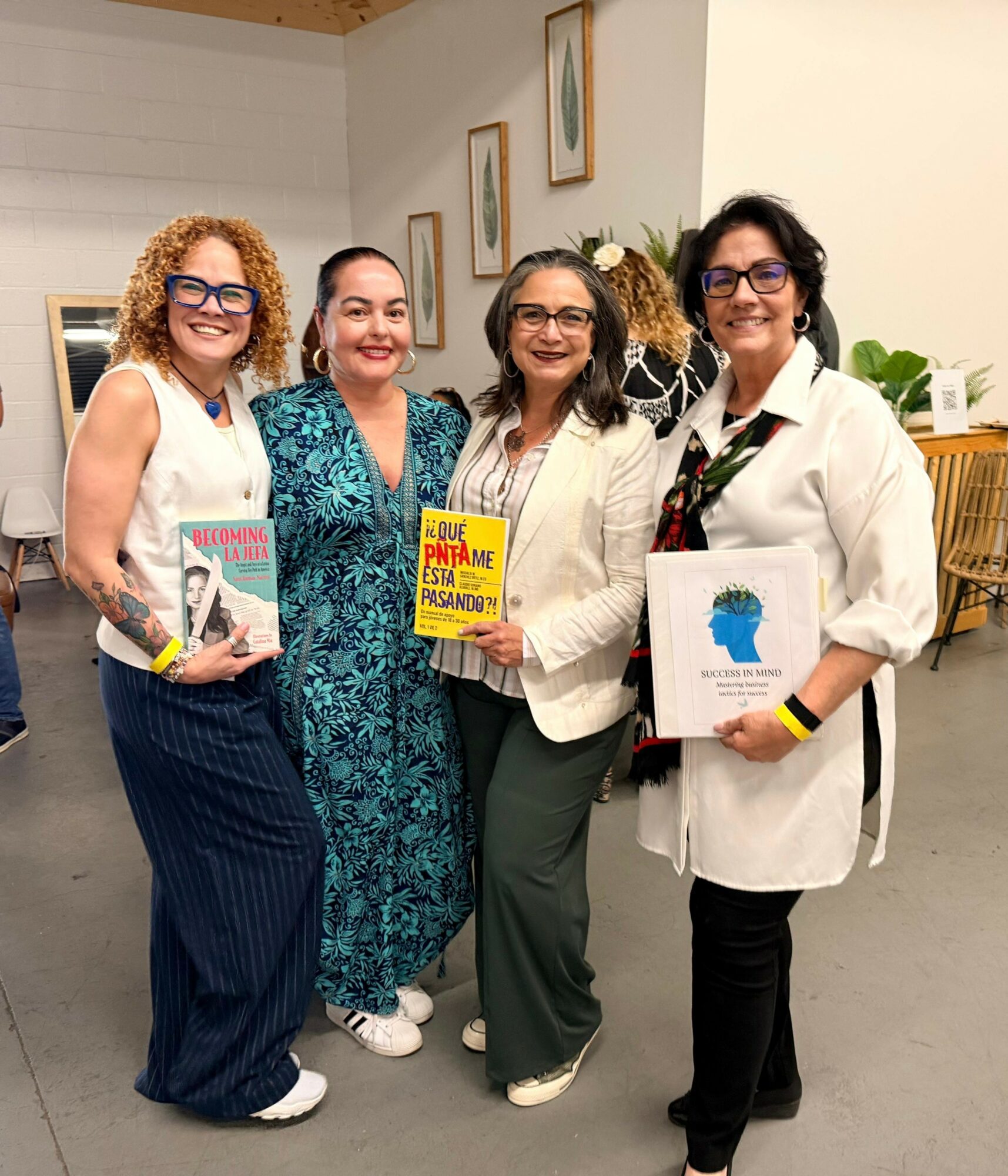 Four women standing indoors, smiling, holding books, with framed artwork on the wall behind them.