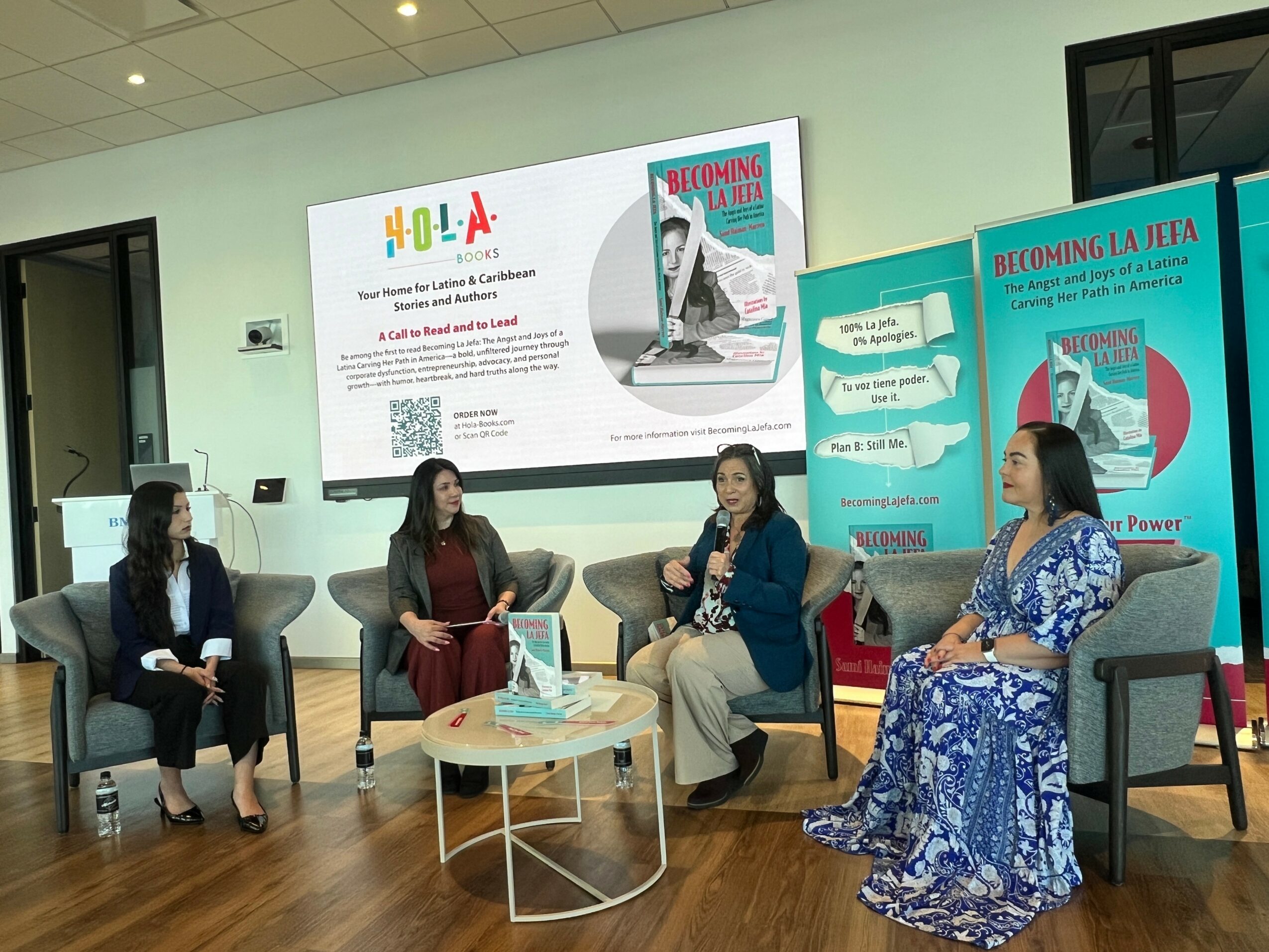 Four women seated in a panel discussion, with a large screen and banners behind them, in a room with wooden floor.
