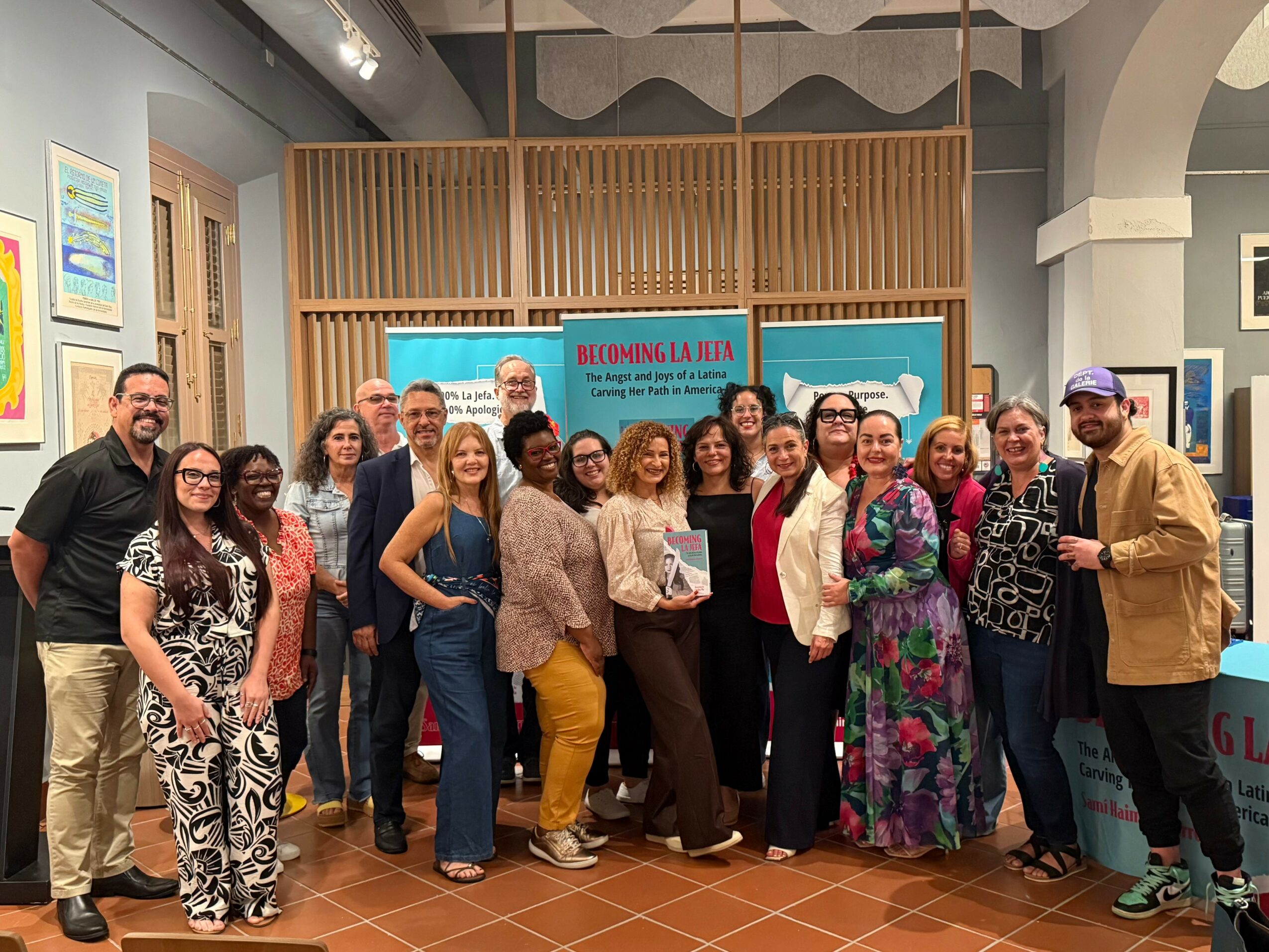 Group of diverse people standing together indoors, some smiling, in front of a screen and wooden partition.