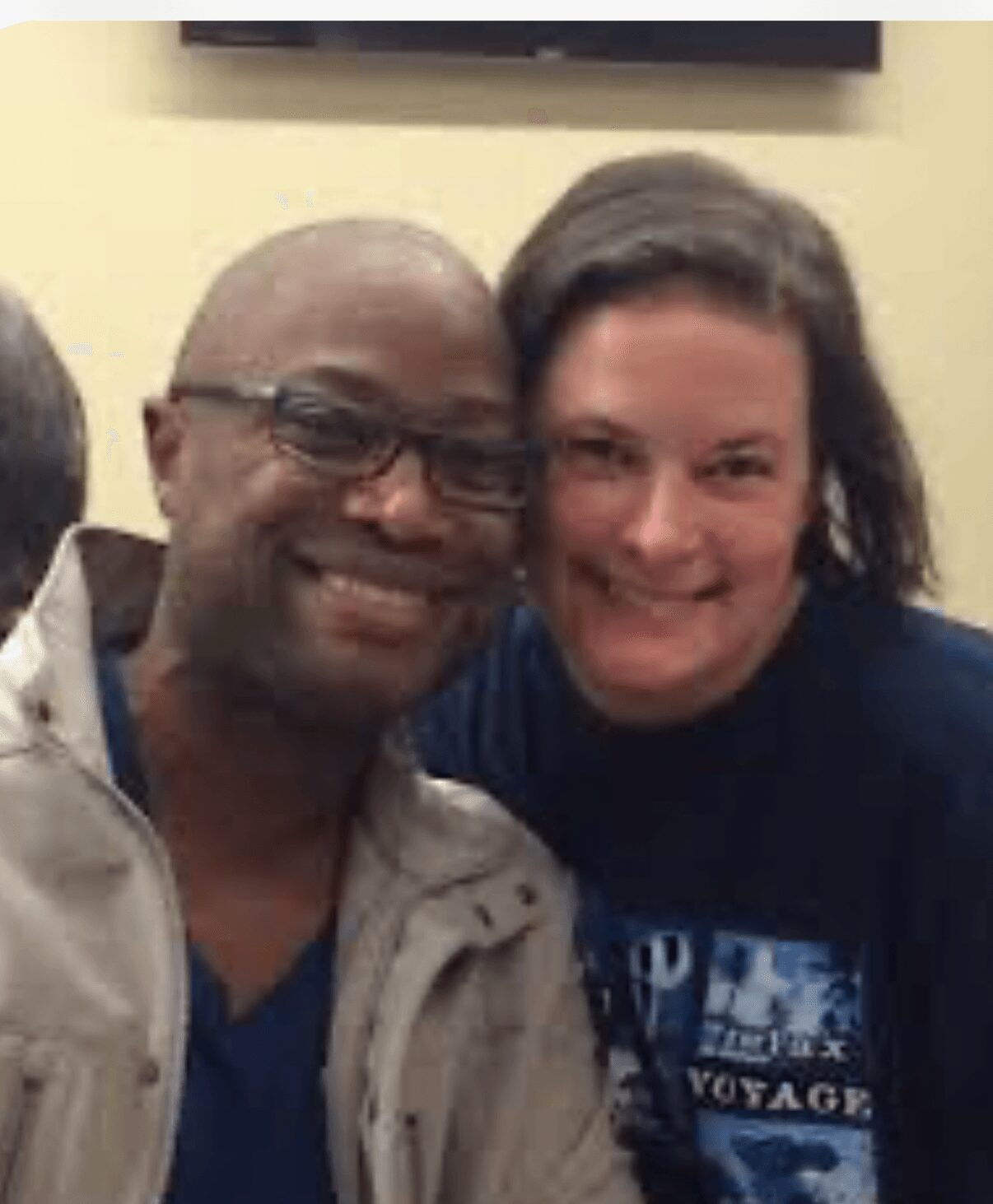 Two people smiling, close together, indoors, with a light-colored wall behind them.