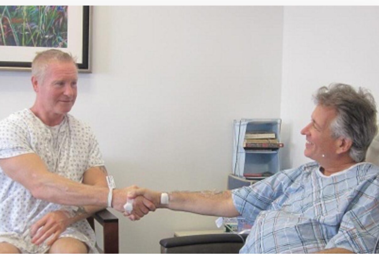 Two men shake hands in a room with a white wall and a framed picture, one seated and one in a chair.