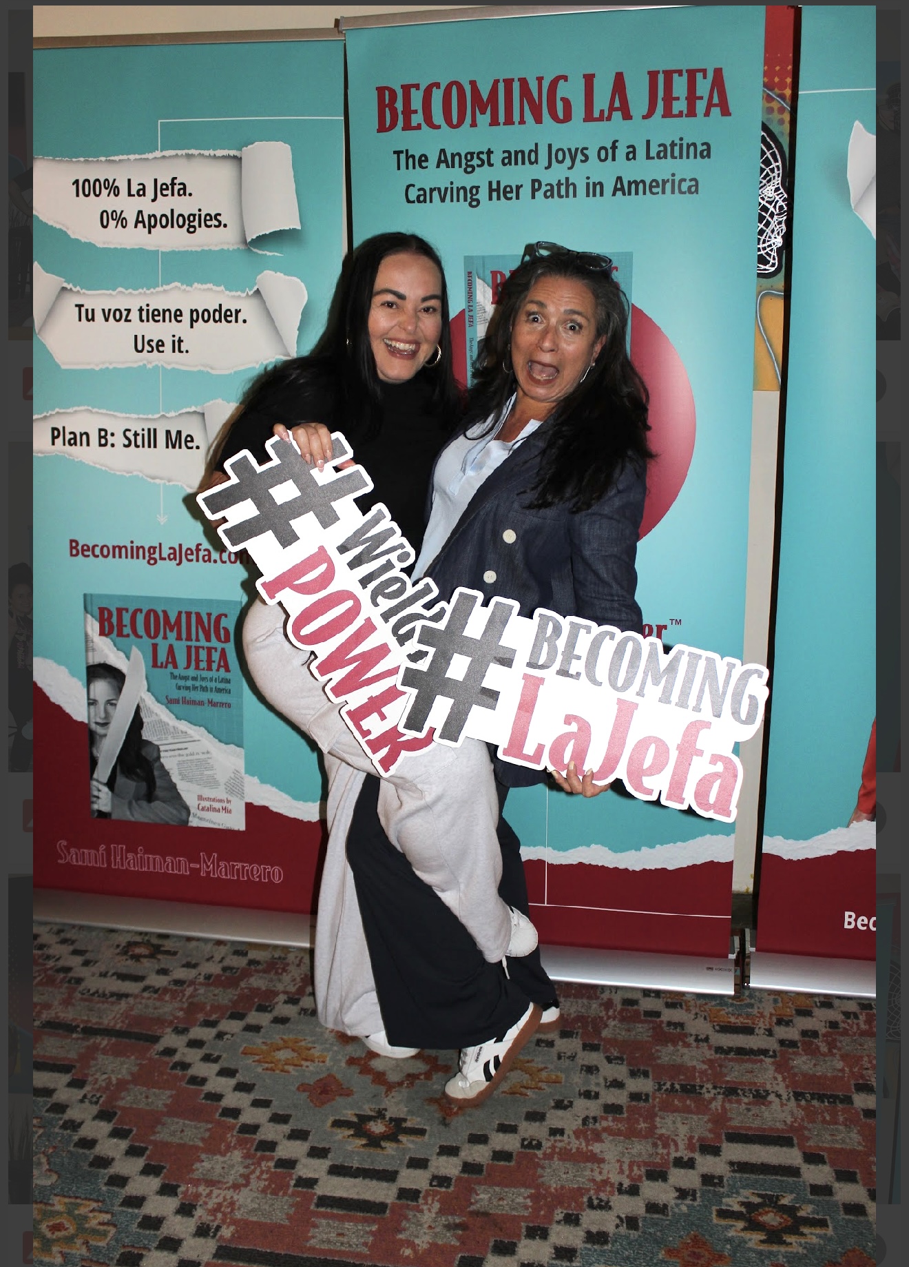 Two women smiling and holding a sign with hashtags, standing in front of a colorful backdrop with text about Latina empowerment.