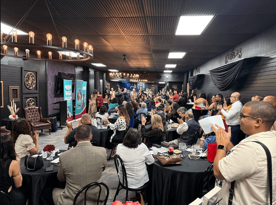 Crowd of people gathered in a large room with tables, some using phones, others talking, with a stage and decorations.