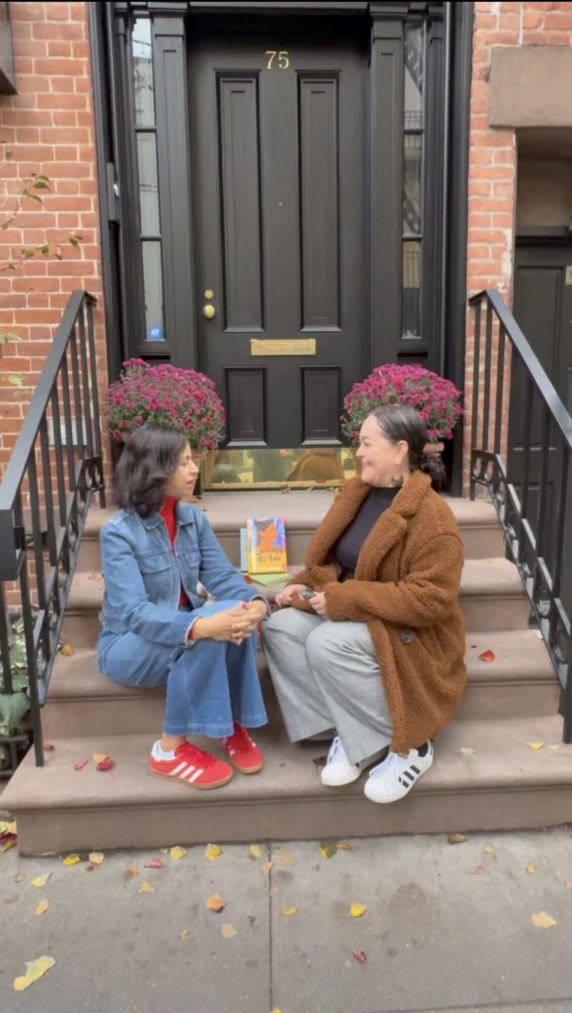 Two women sit on front steps holding hands, with pink flowers in pots and a black door behind them.