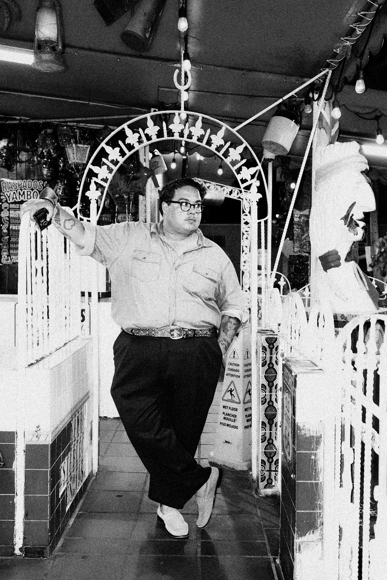 Person standing in front of an archway, pointing with one arm, wearing glasses, a light shirt, and dark pants.