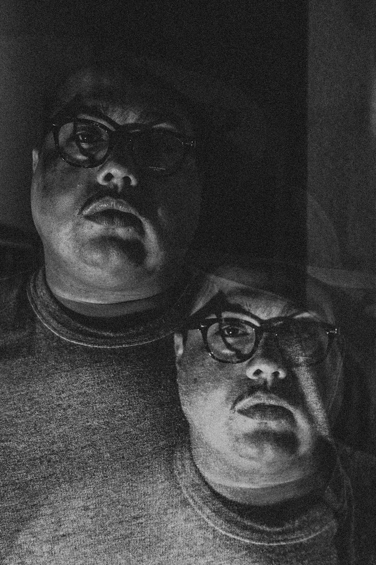 Two people with glasses and similar facial expressions, one larger and one smaller, in a dark setting.