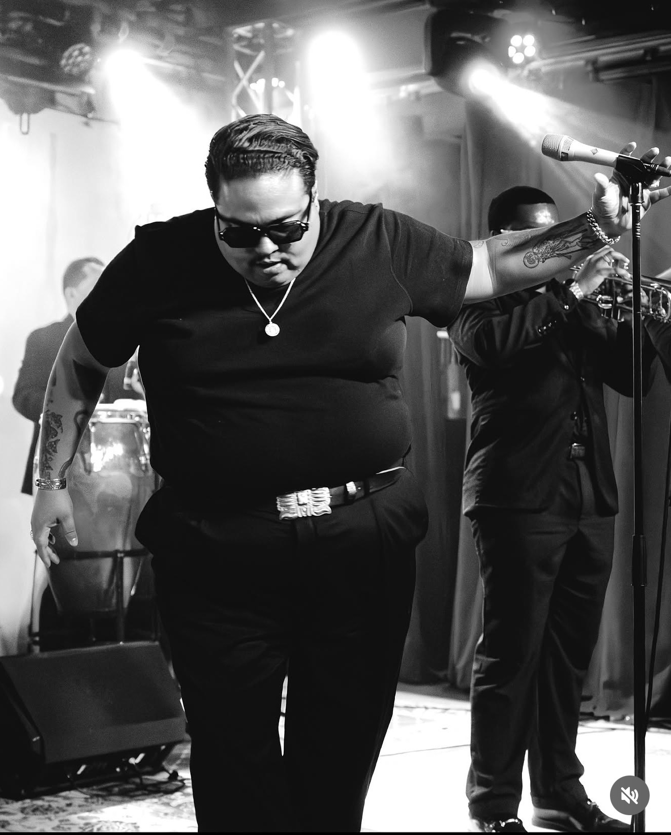 Person with sunglasses and tattoos performing on stage with a microphone, stage lights in background.