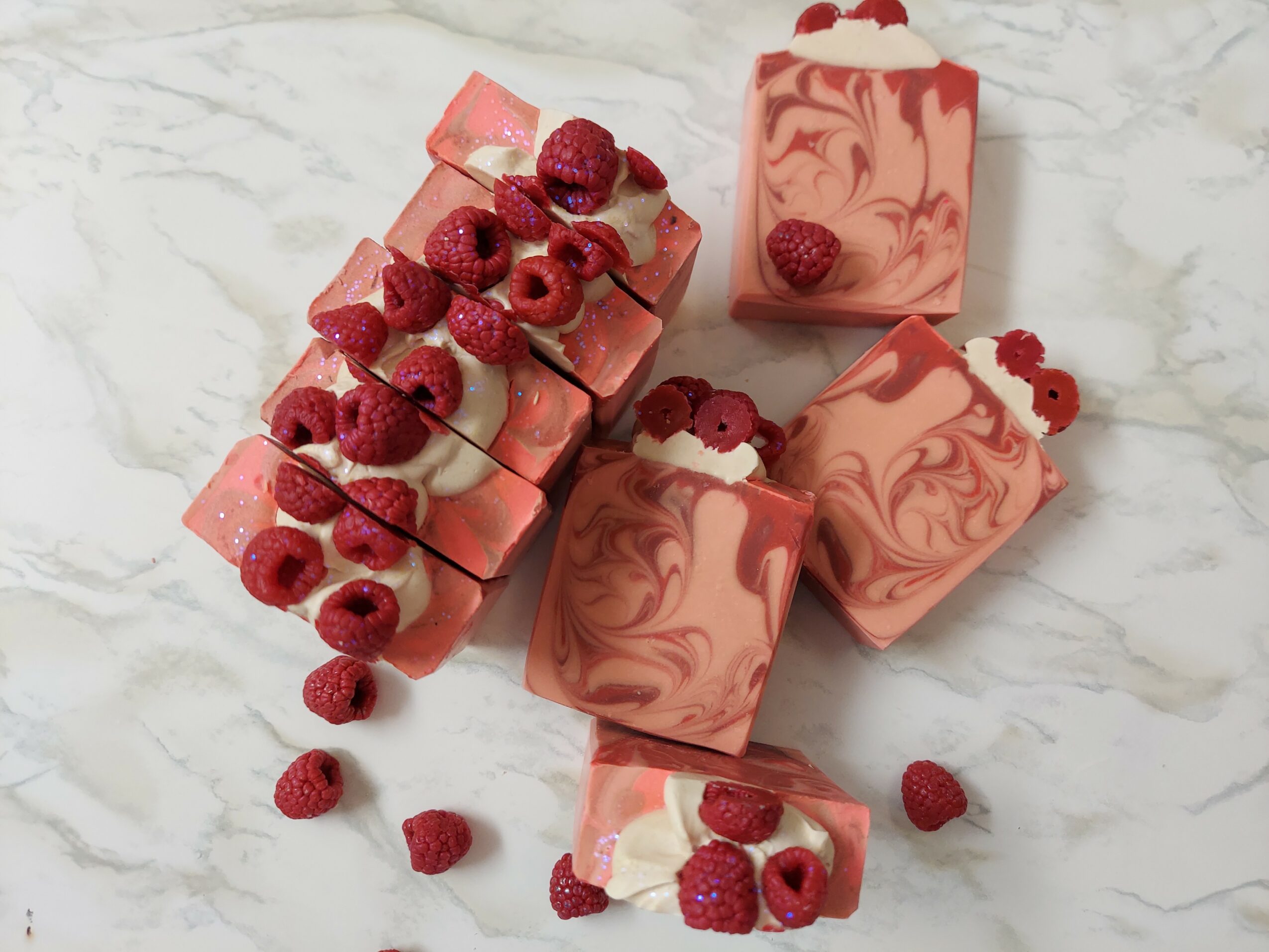 Four pink and white marbled soap bars with raspberries on top, arranged on a marble surface.