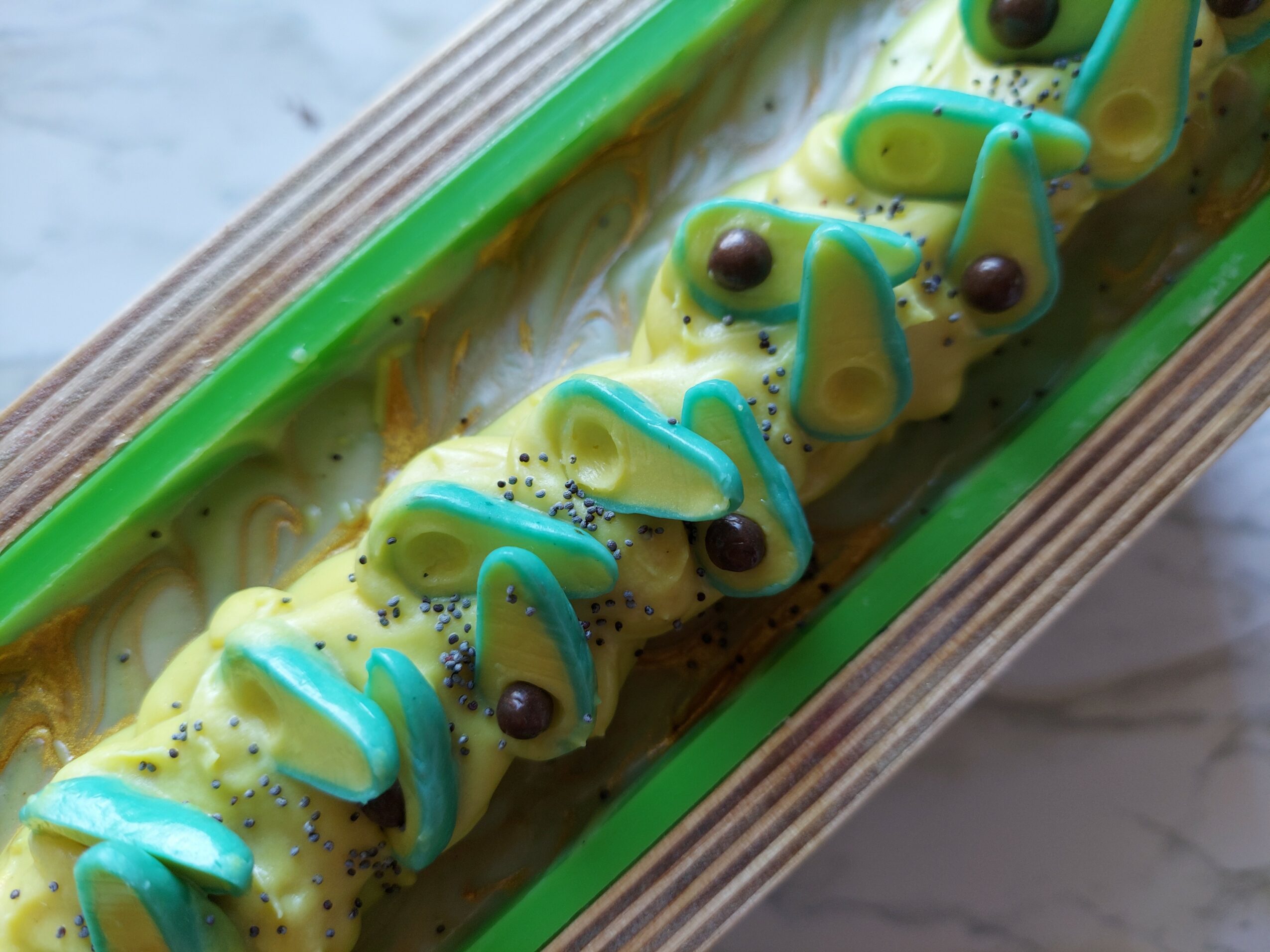 Decorative dessert with yellow and blue icing, small dark toppings, in a green-bordered tray, on a marble surface.