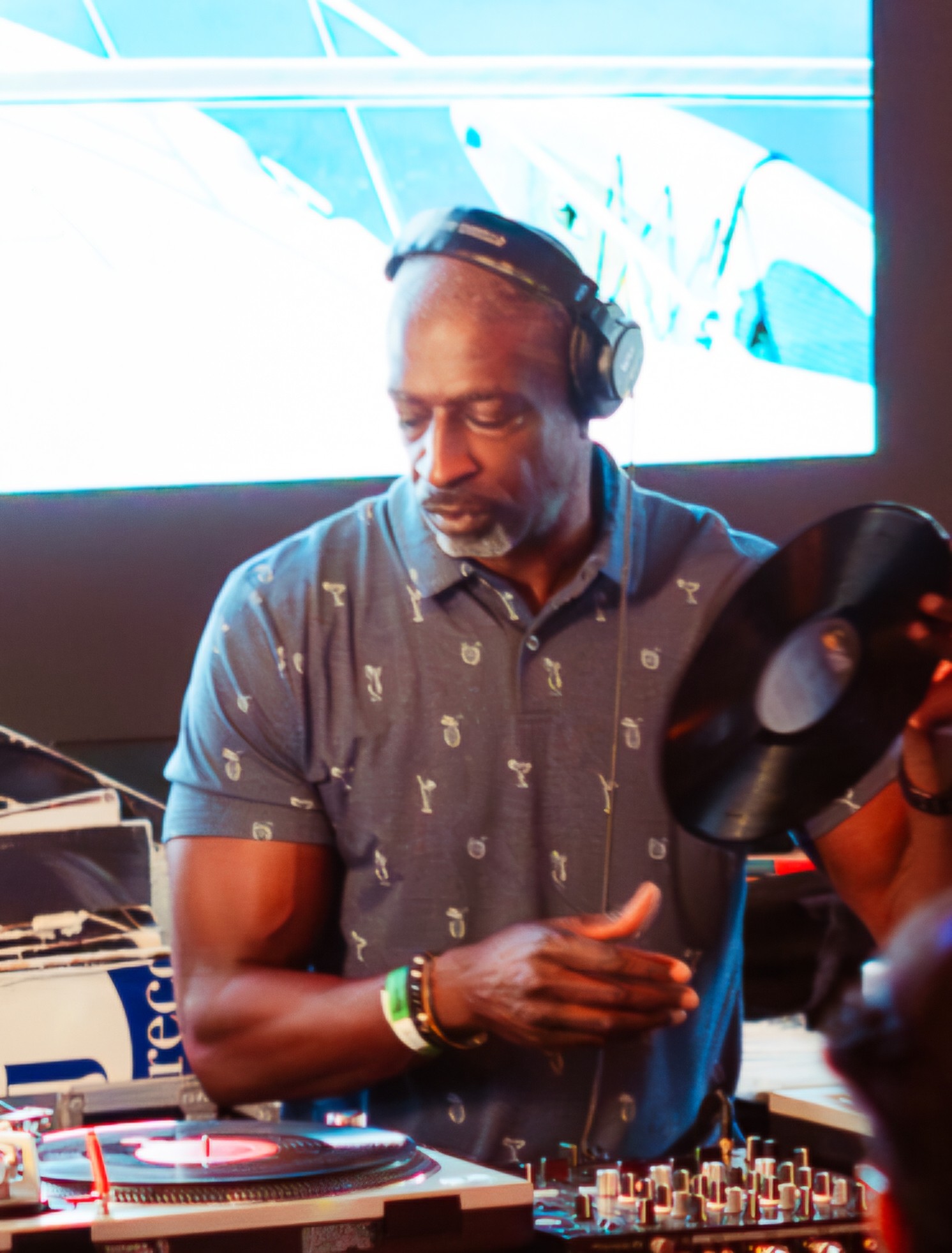 DJ wearing headphones and a patterned shirt, operating turntables with a large screen behind him, in a dimly lit setting.