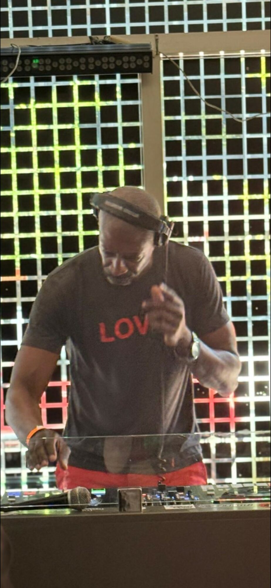 Person with dreadlocks wearing a black shirt with red text, standing in front of a grid background, holding a phone.