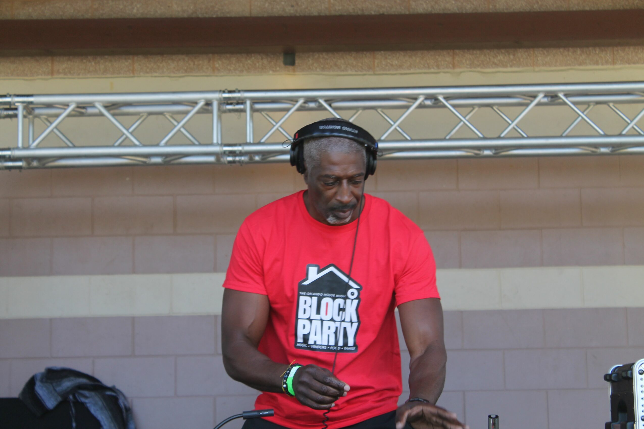 Man wearing headphones and a red T-shirt with 'Block Party' logo, standing outdoors in front of a wall and metal structure.