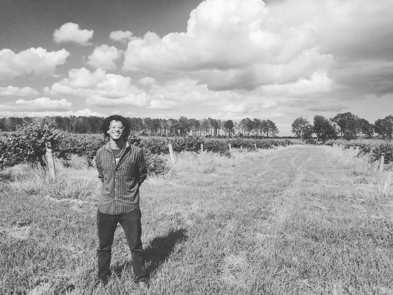 Person standing outdoors in a grassy field with trees and clouds in the background.