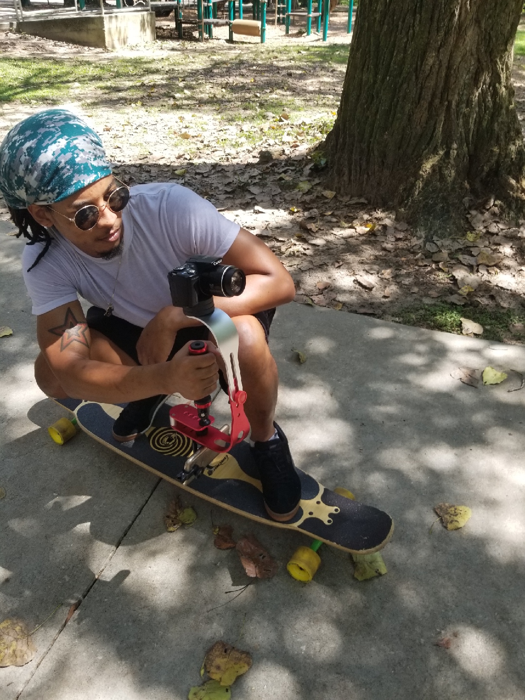 Child wearing sunglasses and a bandana, sitting on a skateboard under a tree in a park, holding a camera.