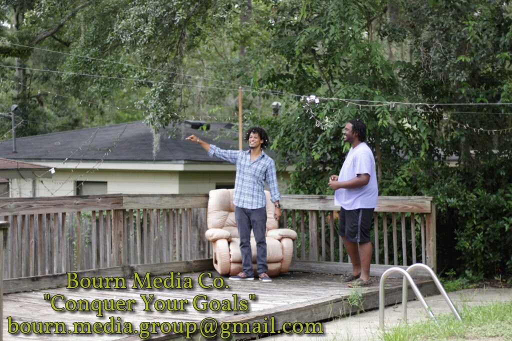 Two men standing on a wooden deck outdoors, one gesturing with his arm, surrounded by trees and a fence.