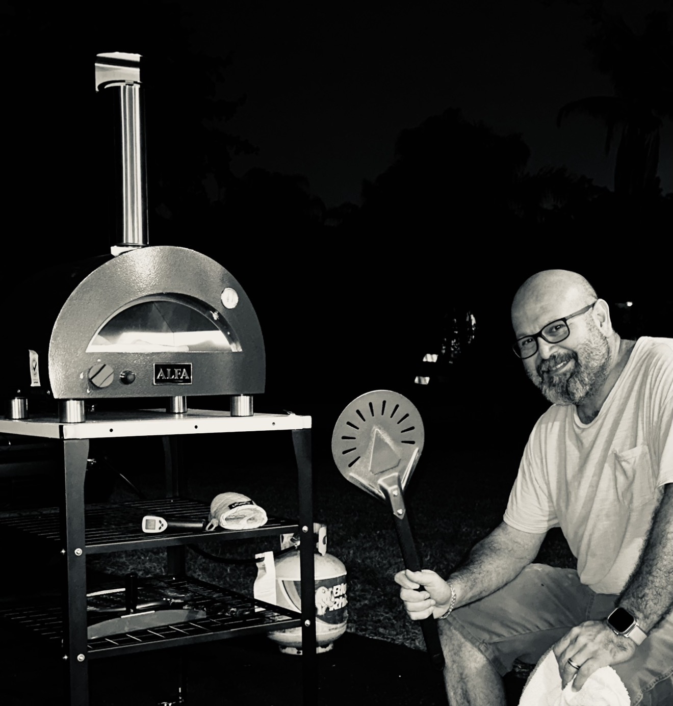 Man with glasses and beard sitting outdoors at night, holding a pizza peel, with a pizza oven nearby.
