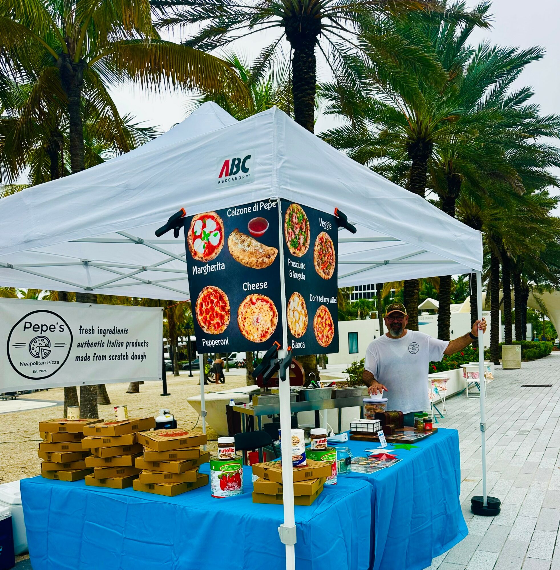 Food stand with a white canopy, blue tablecloth, and a person smiling, surrounded by palm trees.