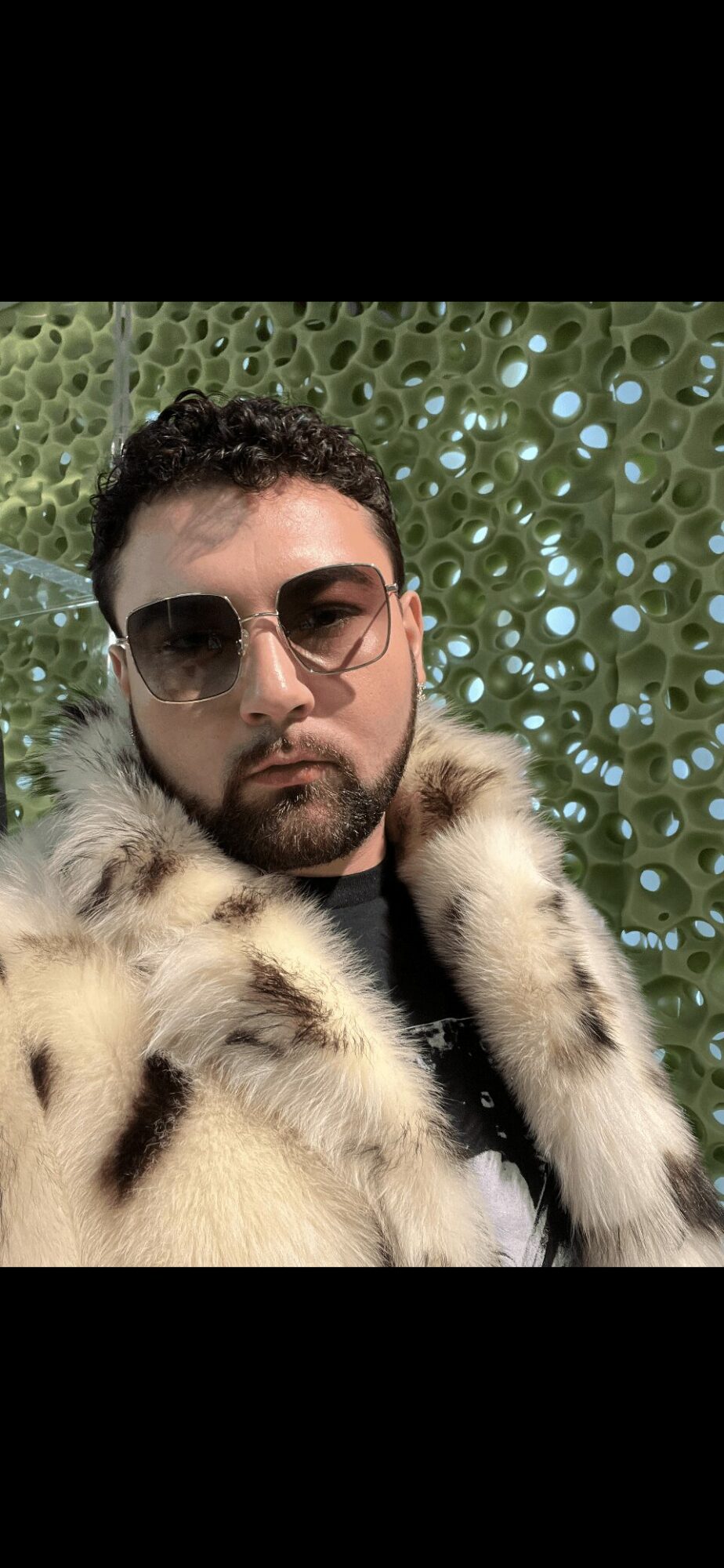 Man with sunglasses and fur coat in front of a perforated green background.