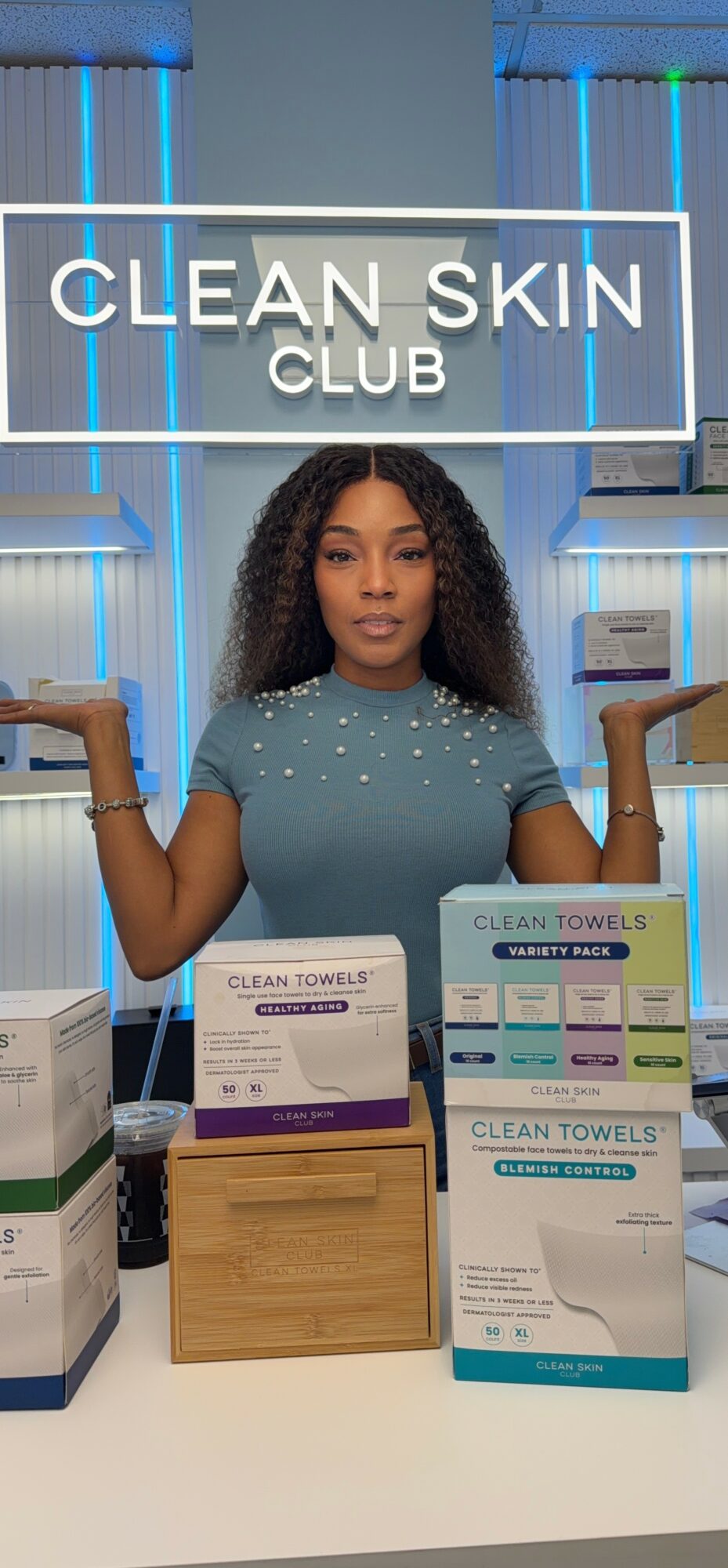 Woman with curly hair standing behind a counter with skincare products, in front of a sign reading 'CLEAN SKIN CLUB'.
