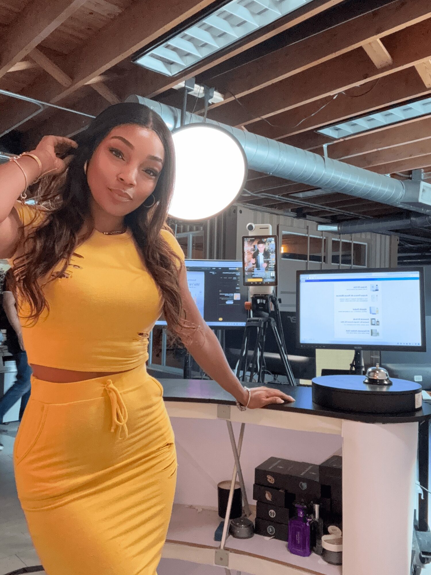 Woman in yellow dress standing in a studio with cameras and monitors, holding a microphone, with a large light overhead.