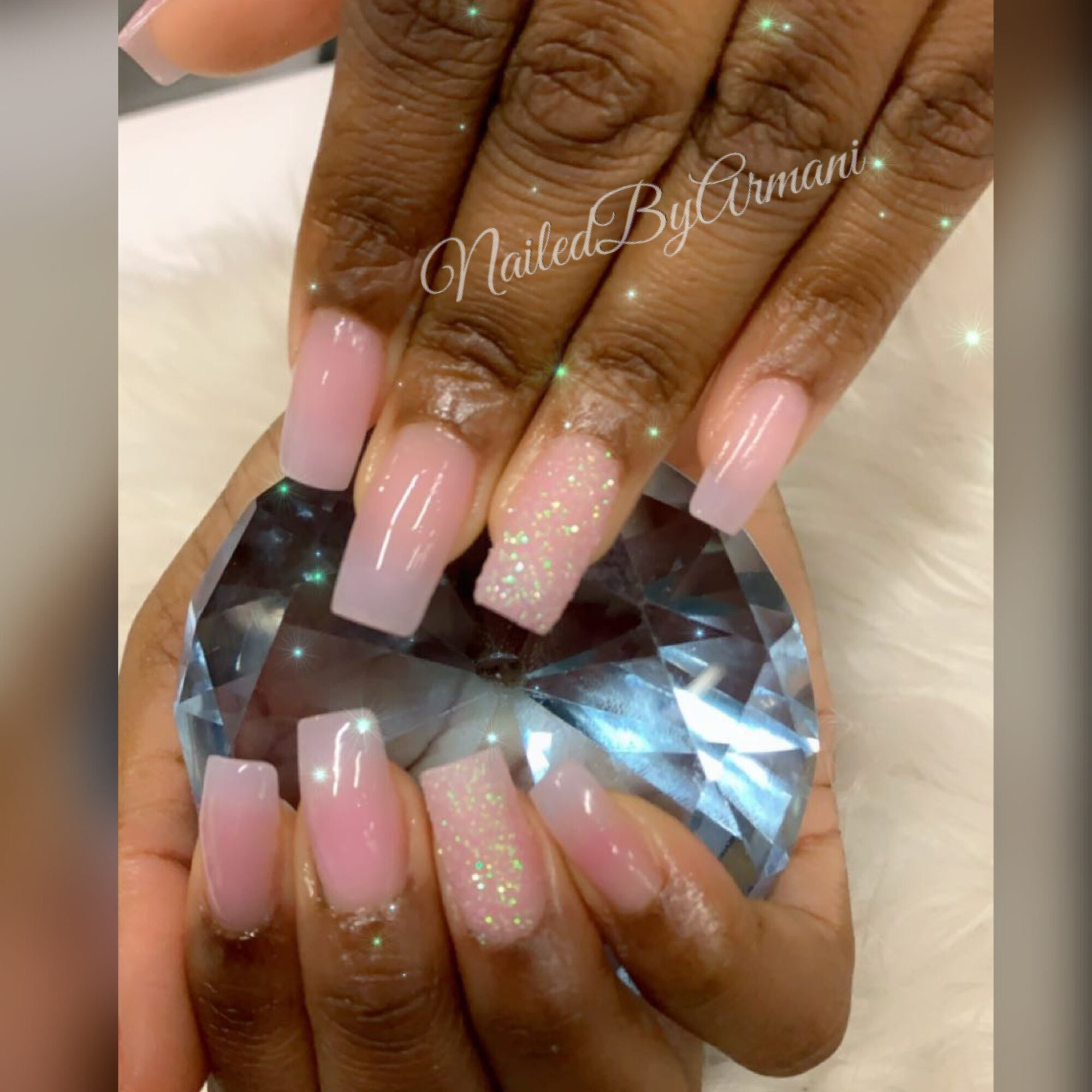 Hand with long pink and glittery nails holding a large diamond-shaped crystal, background is neutral.