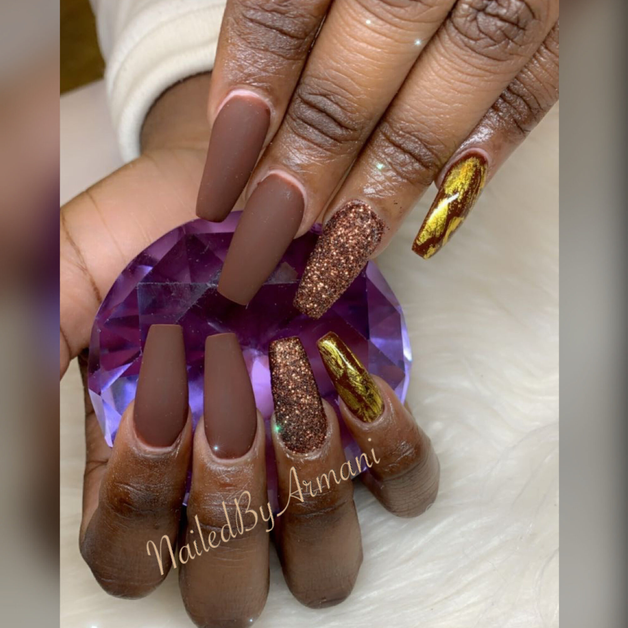 Close-up of hands with manicured nails, featuring matte brown, glitter, and gold designs, holding a purple gemstone. Word watermark reads 'NailedByAmani'.