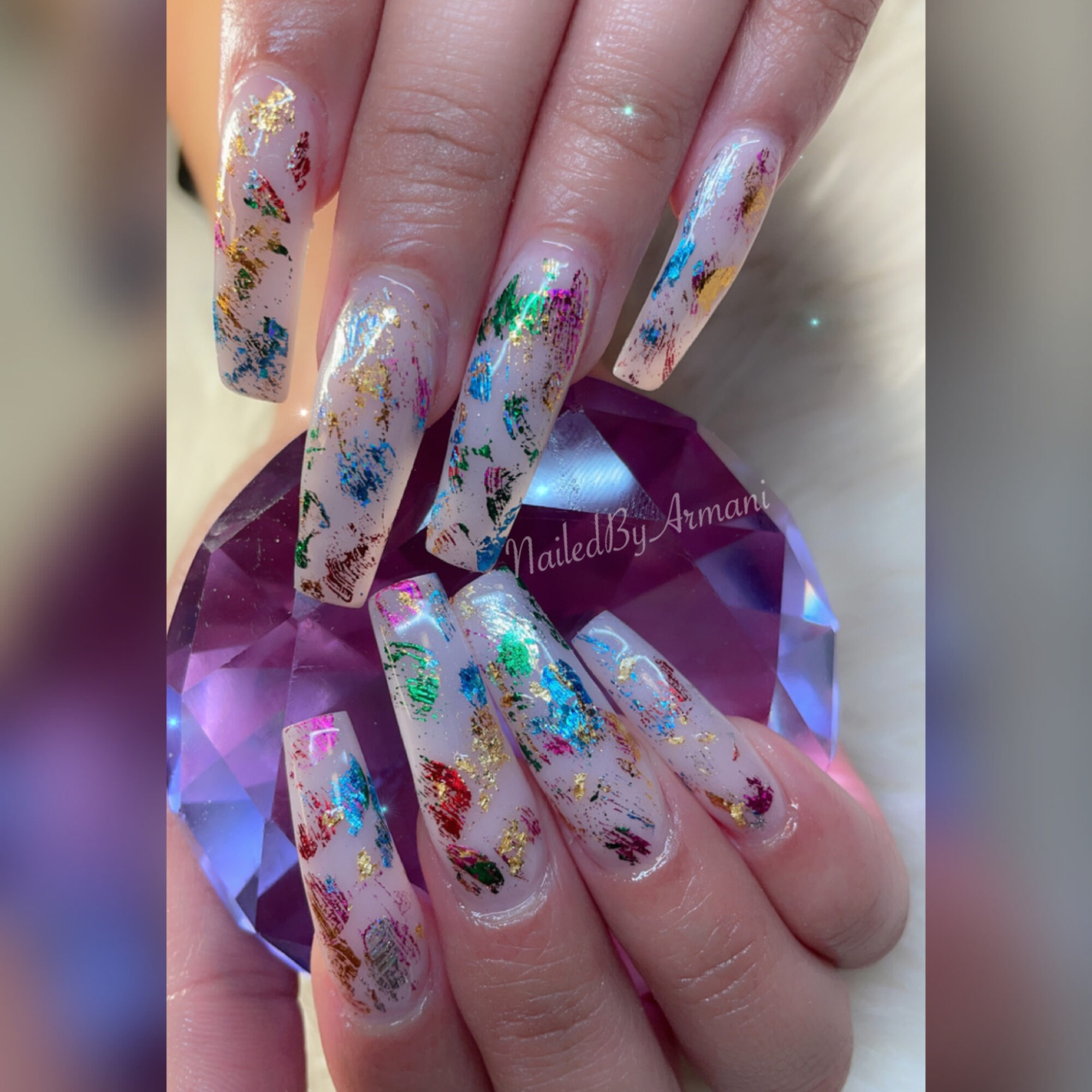 Hand with long, decorated nails resting on a purple gemstone, nails have colorful abstract designs with glitter and metallic accents.