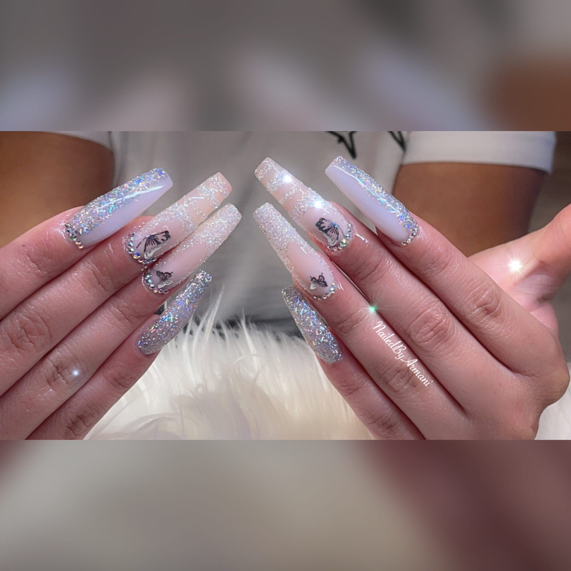 Person's hands with long, glittery, decorated nails, holding a white furry surface, background shows part of person's arms and shirt.