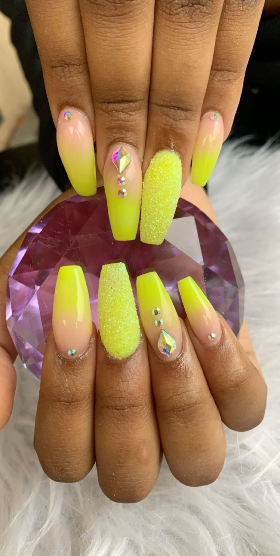 Close-up of hands with long, yellow, textured and decorated nails holding a pink gemstone, with a white furry background.