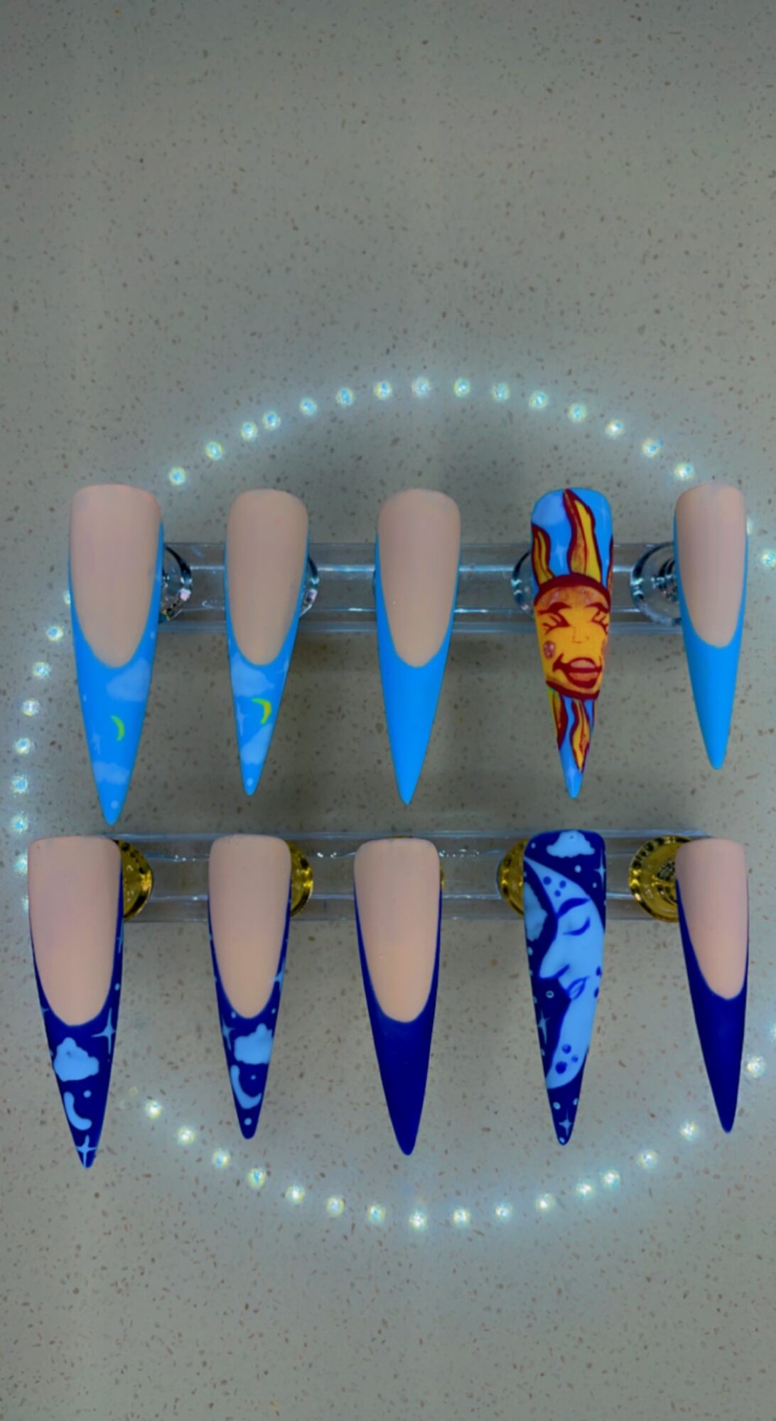 Two rows of artificial nails with blue and beige designs, one with a colorful fish design, on a gray surface.