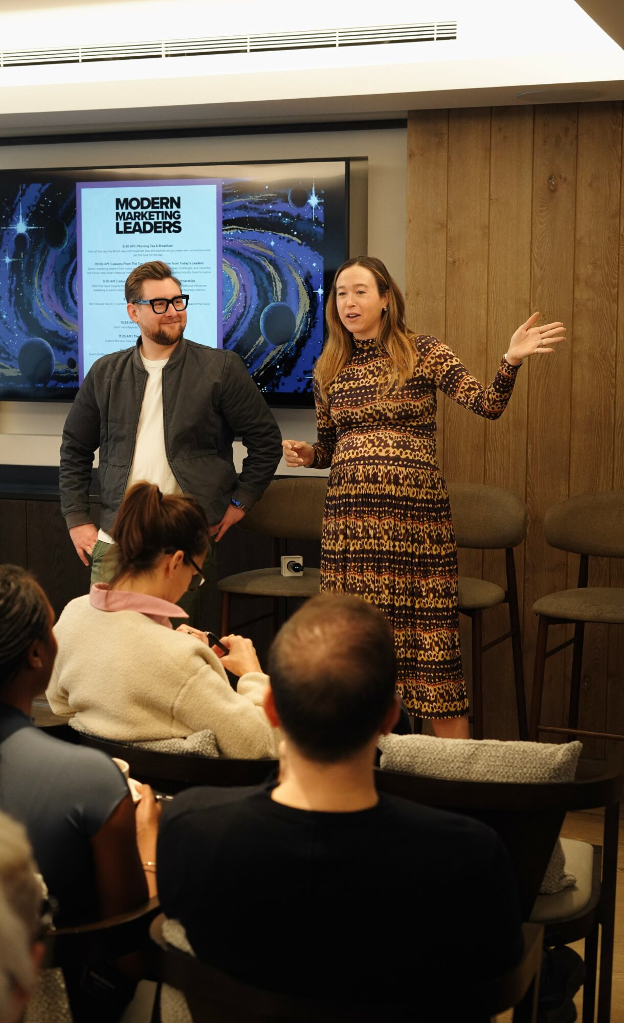 Two people stand in front of an audience, one speaking and gesturing, the other listening, with a presentation screen behind them.