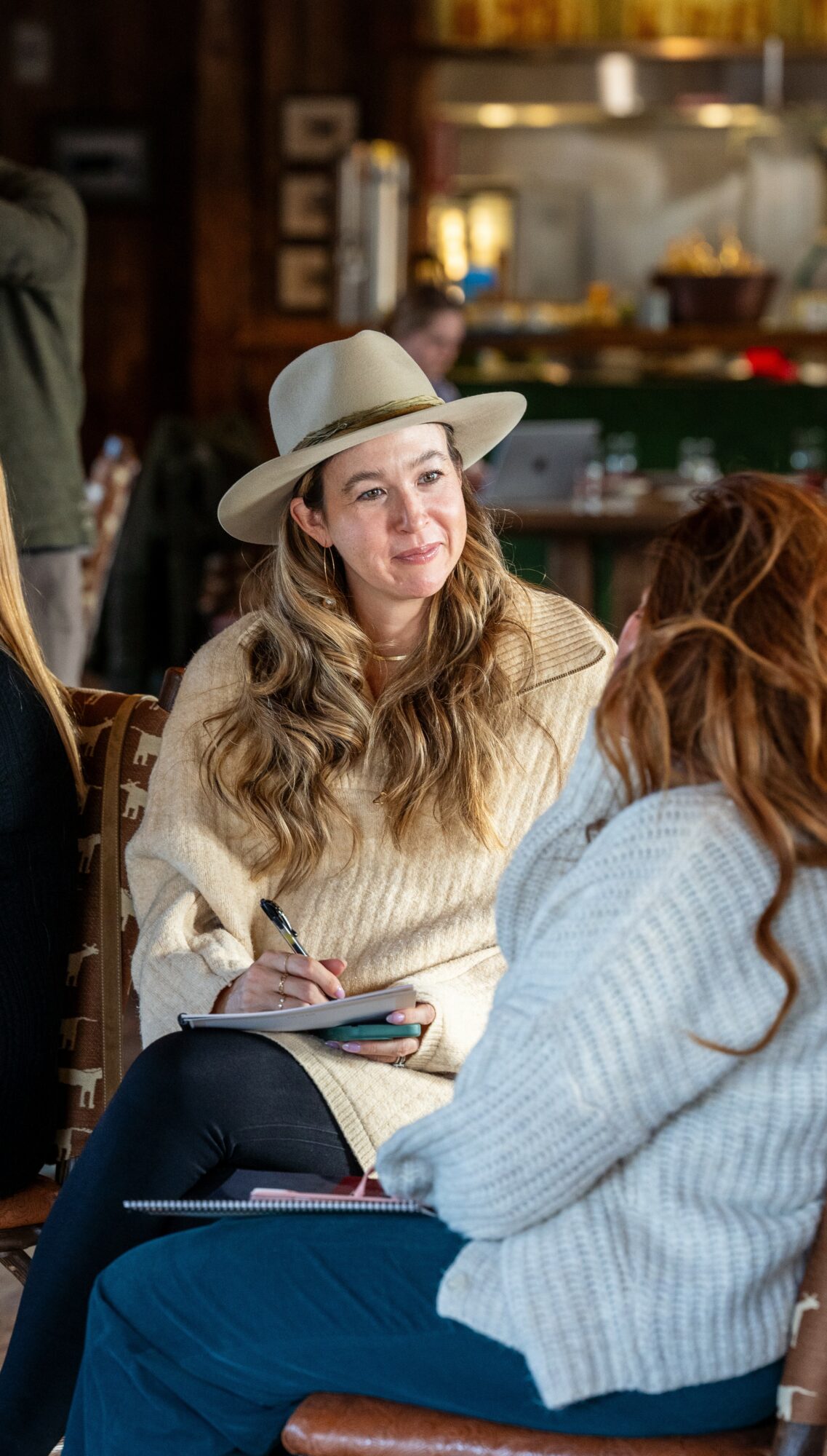 Woman with long hair wearing a hat and cream sweater, sitting and writing in a notebook, in a cozy indoor setting.