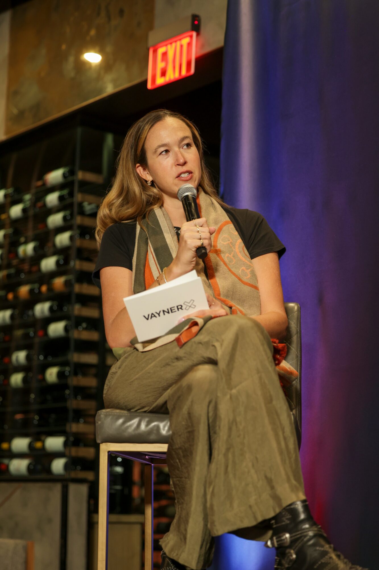 Woman speaking into microphone, seated on stage with wine bottles in background, illuminated by stage lighting.