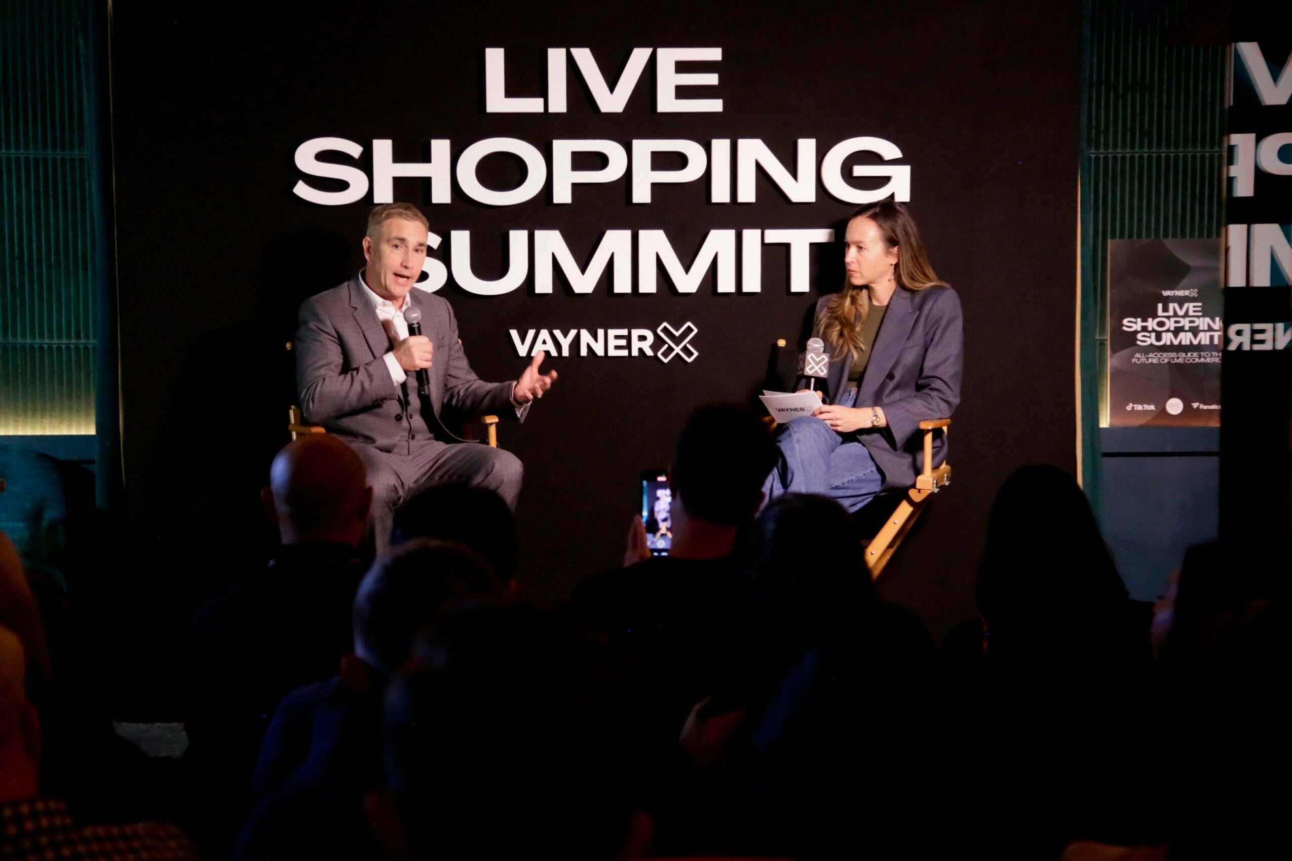 Two people seated on stage in front of a large screen with text 'LIVE SHOPPING SUMMIT' and 'VAYNER'. Audience members are visible in the foreground.