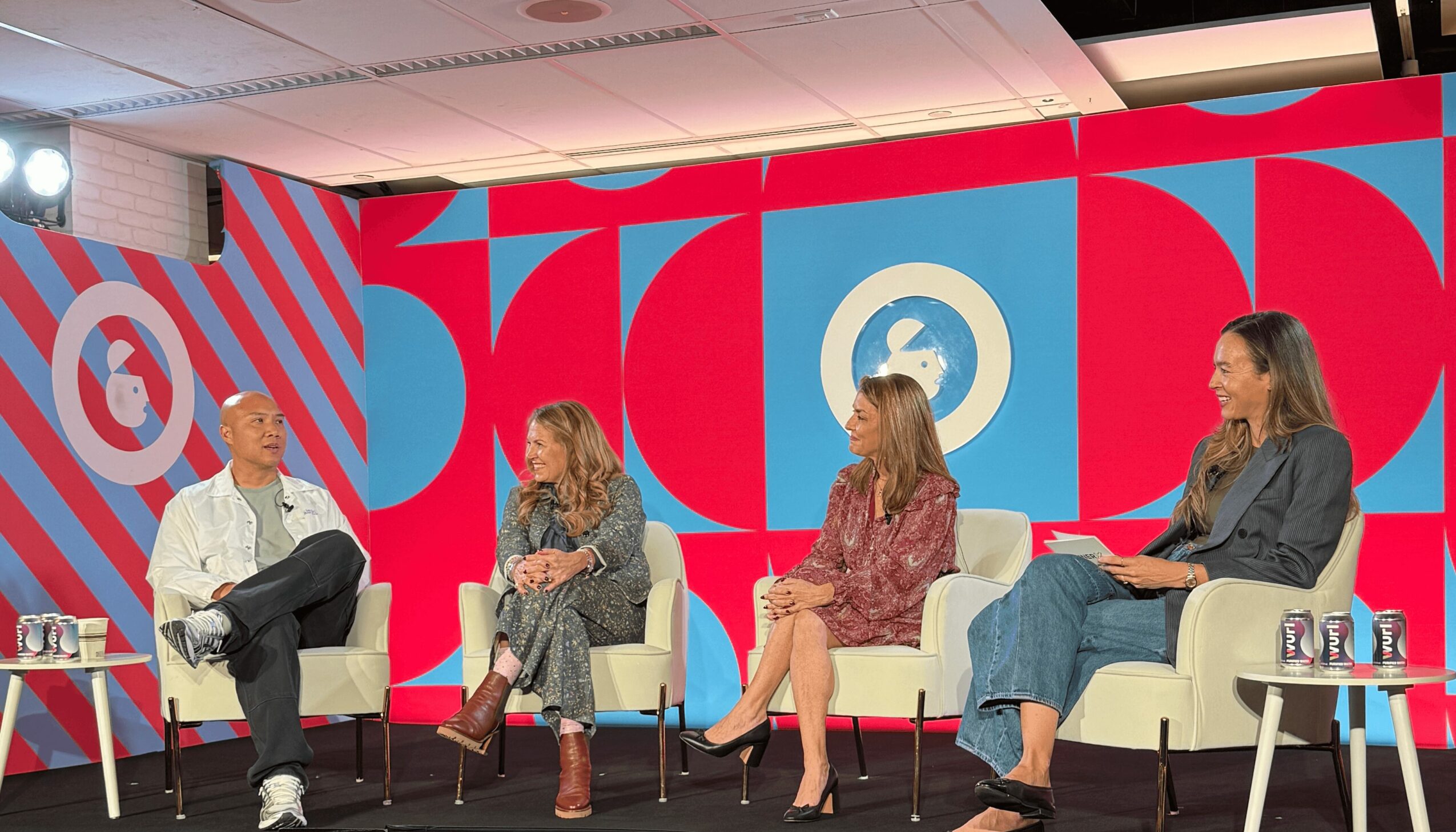 Four women seated on stage in discussion, with a colorful backdrop featuring large red and blue shapes and a logo.