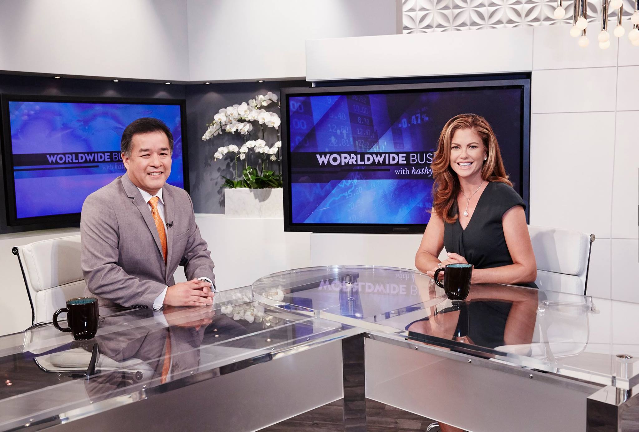 Two news anchors sit at a transparent desk in a studio with screens behind them displaying 'Worldwide Business.'