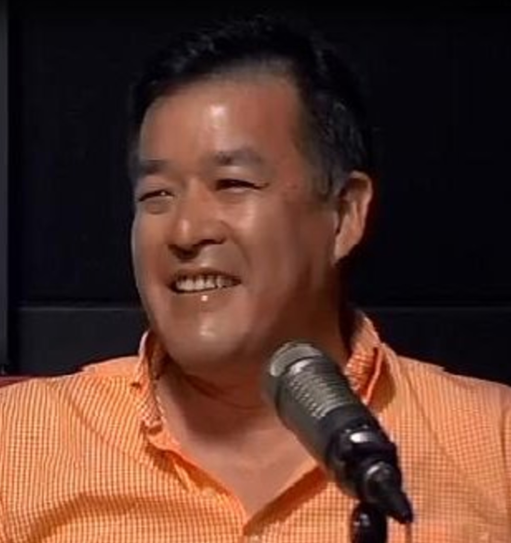 Man smiling, wearing an orange shirt, speaking into a microphone, dark background.