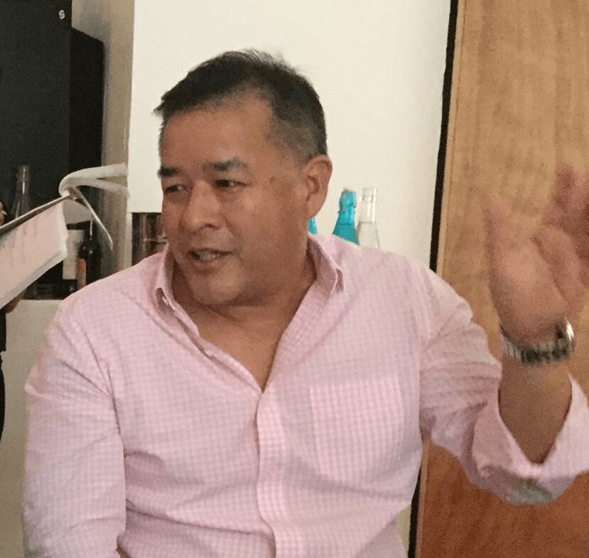 Man with short dark hair wearing a pink checkered shirt, sitting indoors, gesturing with his right hand, smiling slightly.