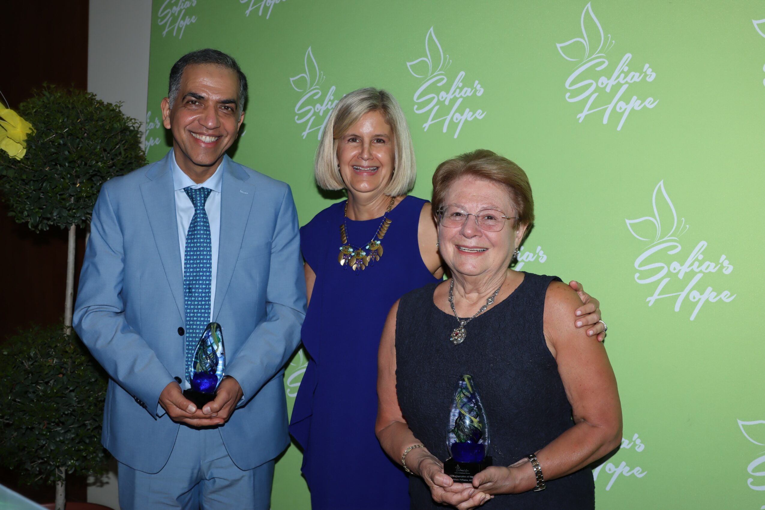 Three people smiling, standing in front of a green backdrop with white logo and text, holding awards, with a potted plant nearby.