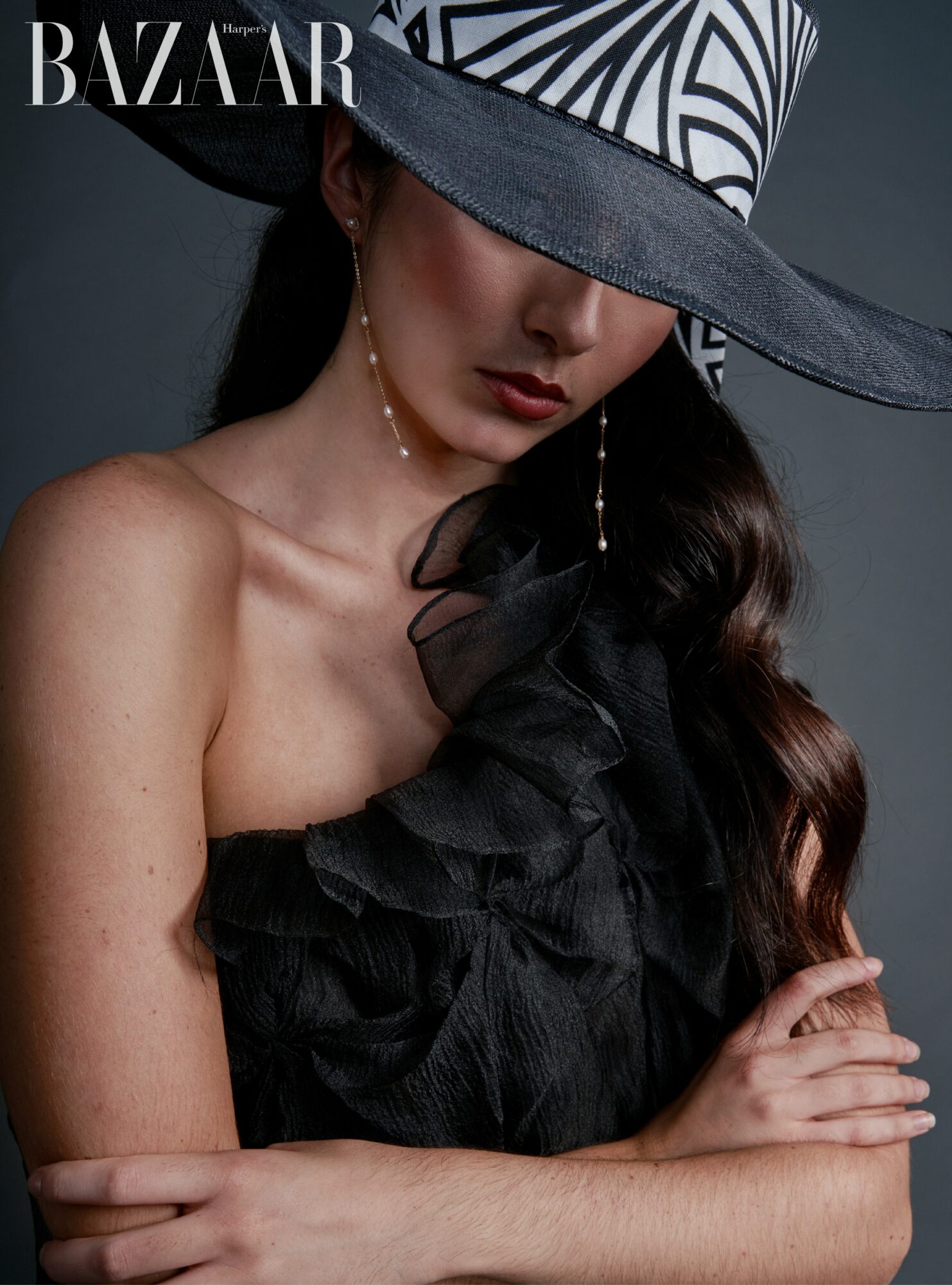 Woman wearing a large patterned hat and black ruffled dress, with long wavy hair and a neutral expression.