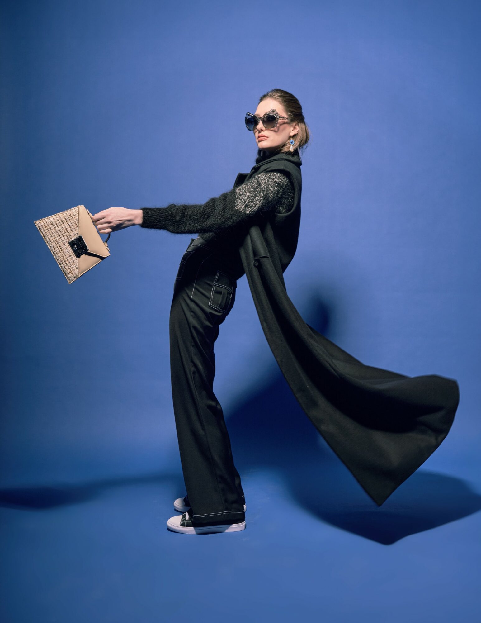 Woman in black outfit holding a woven handbag, wearing sunglasses, standing against a blue background with a flowing cape.