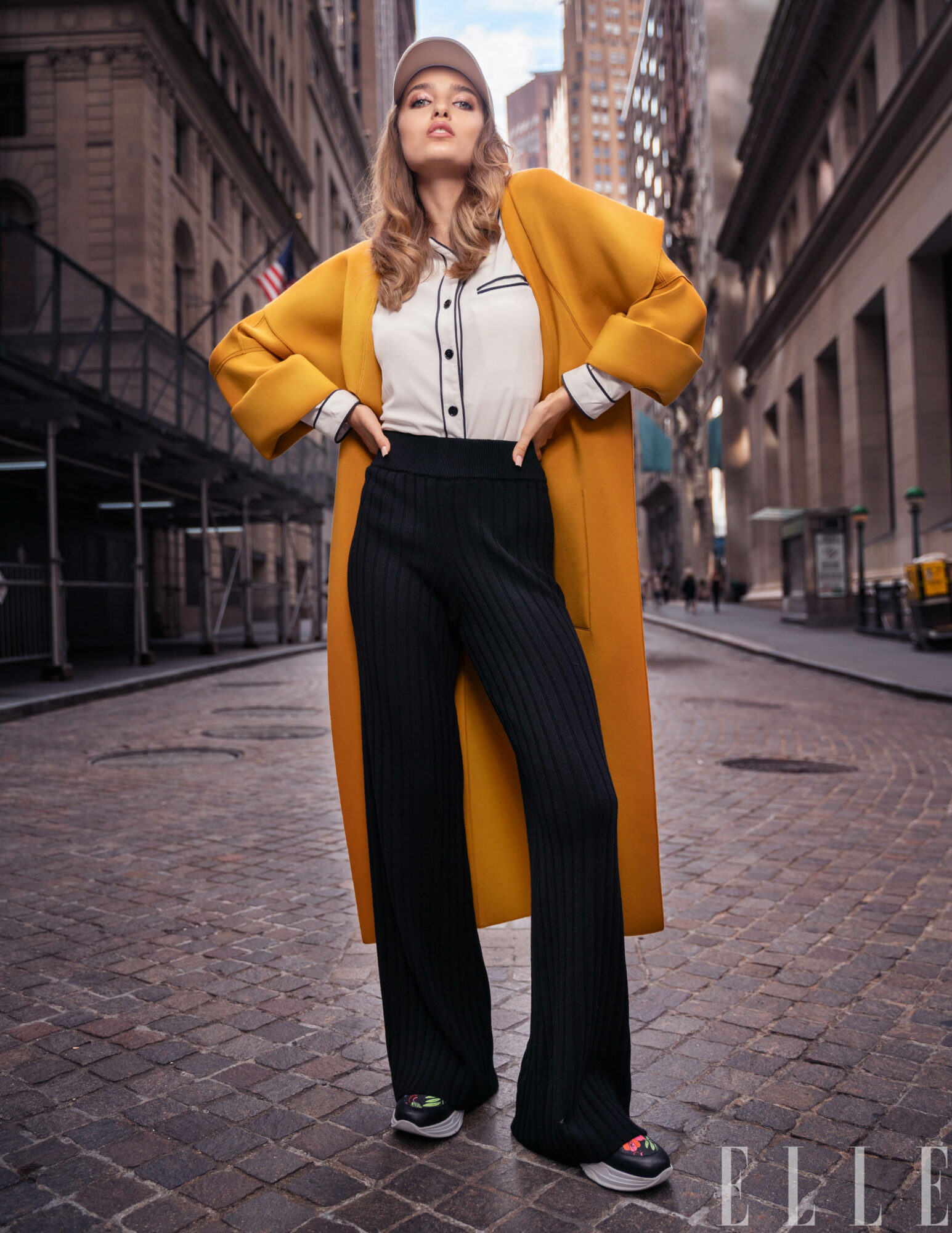 Woman in a yellow coat, white shirt, and black pants standing on a city street with tall buildings.