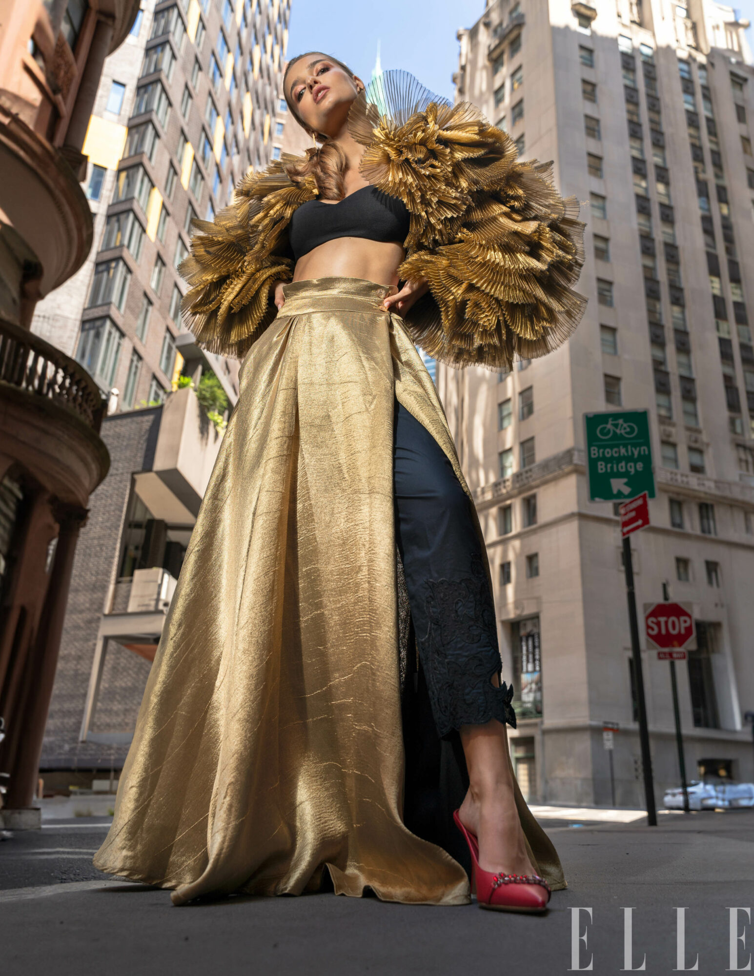 Woman in gold and black outfit with a large gold cape, standing on city street with tall buildings around.