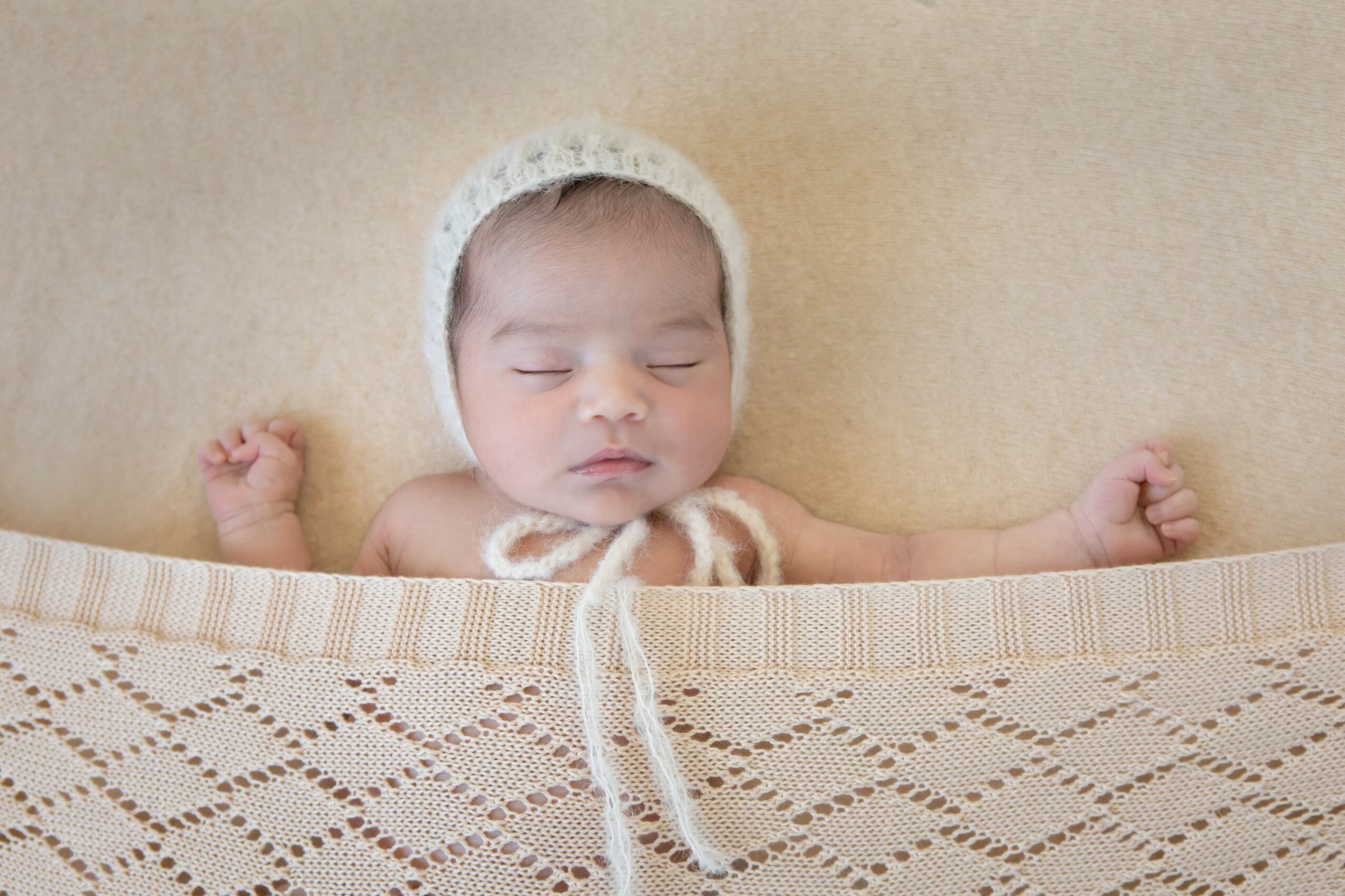 Baby with closed eyes wearing a knit hat, lying on a beige surface, partially covered by a knitted blanket.