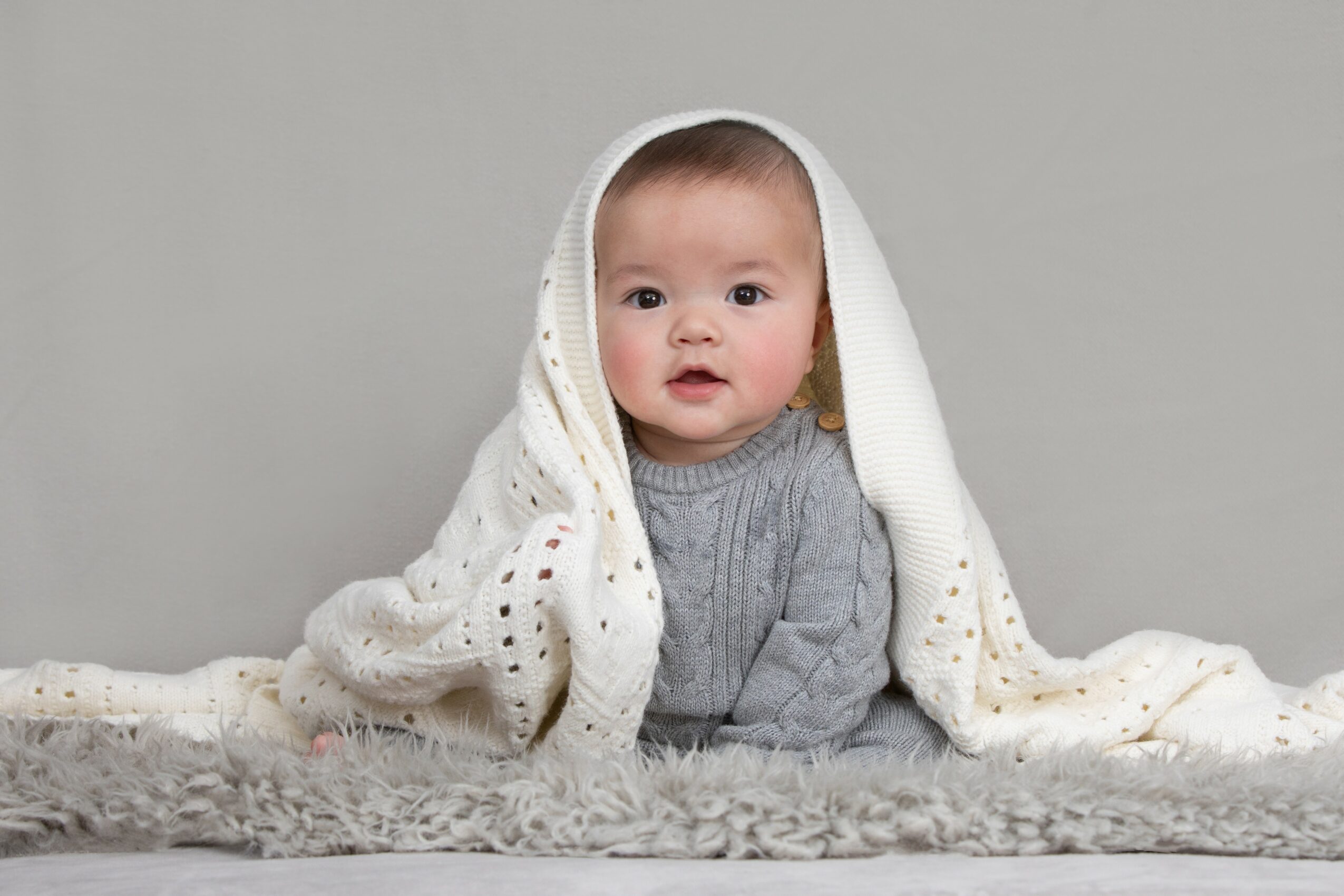 Baby with a gray sweater, covered with a white blanket, sitting on a soft surface against a plain background.