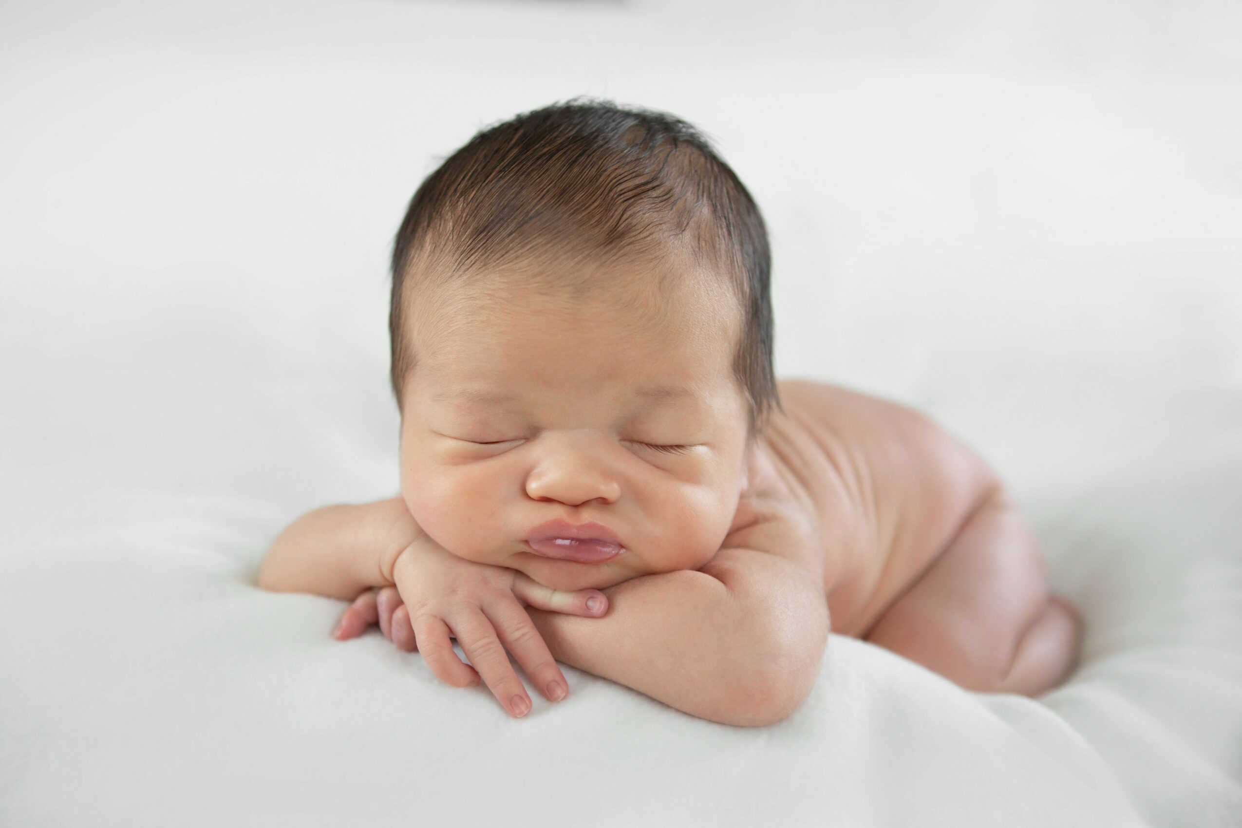 Newborn baby lying on stomach with head resting on hands, eyes closed, on a soft white surface.