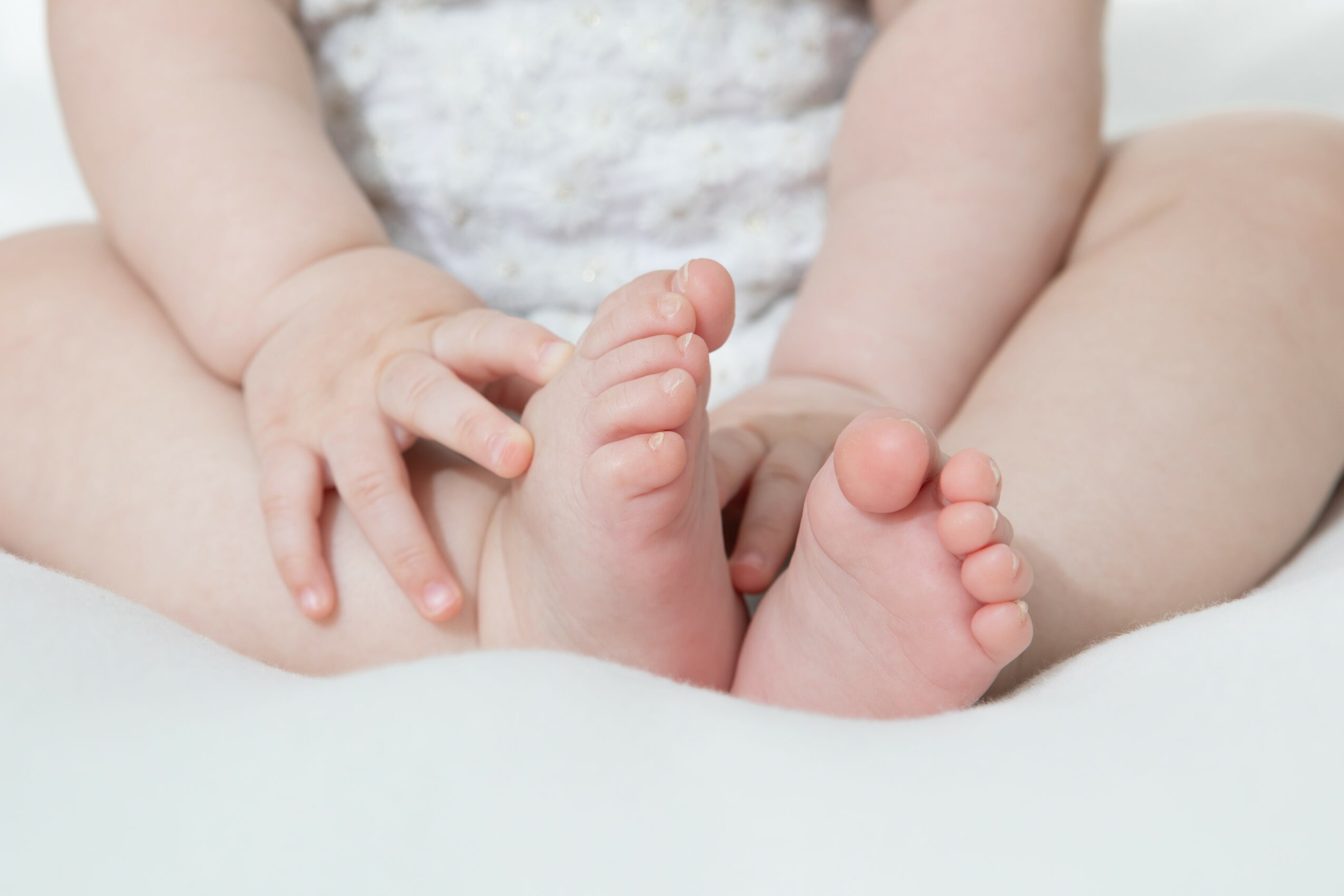 Close-up of a baby's feet and hands, with the baby sitting on a soft surface, wearing a light-colored outfit.