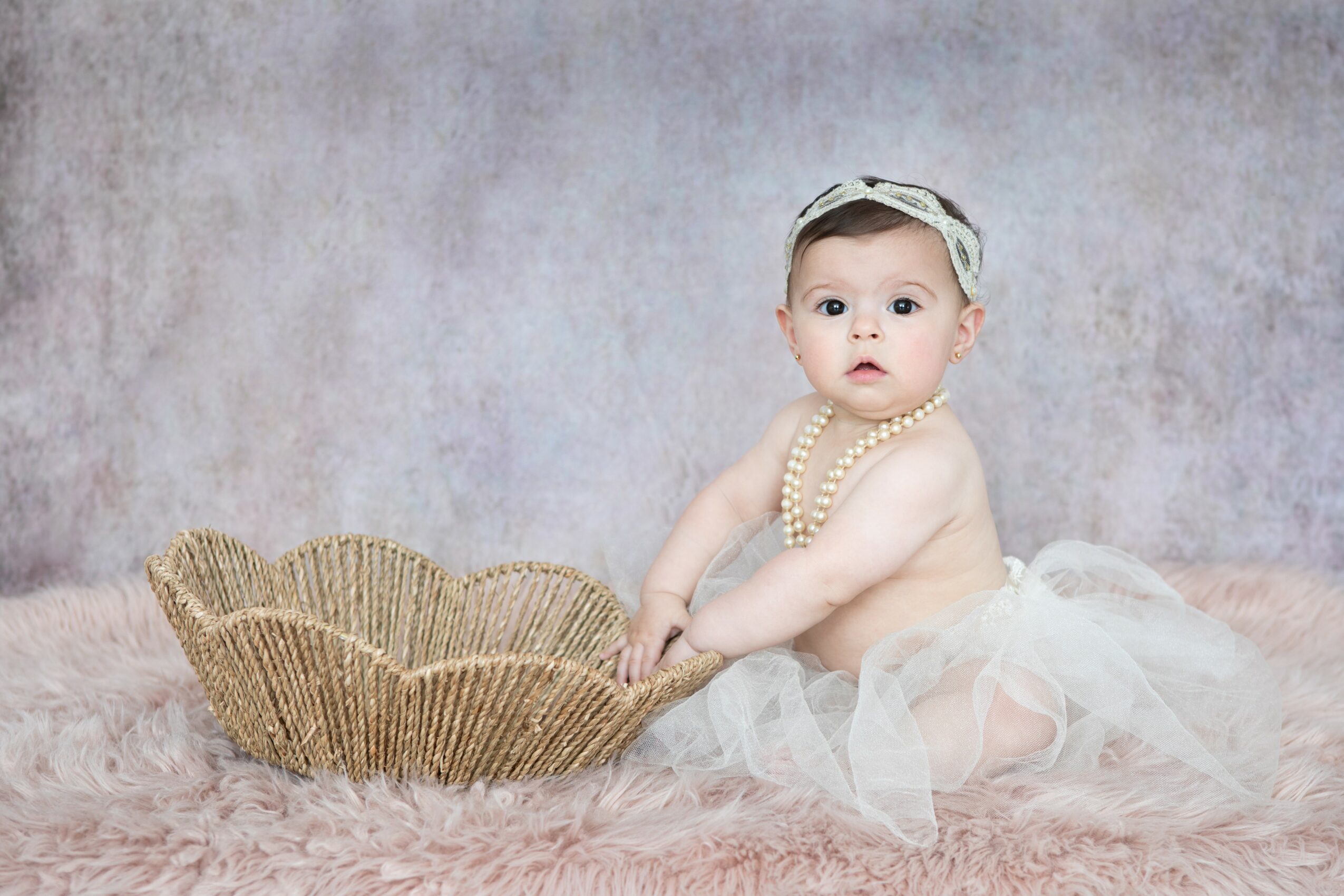 Baby girl sitting on pink fluffy surface with a wicker basket, wearing a tulle skirt, pearl necklace, and headband, looking at camera