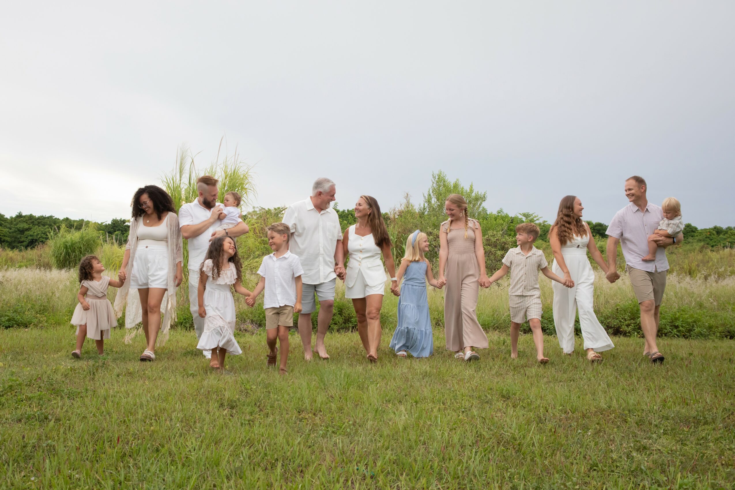 Family of fifteen walking outdoors on grass, holding hands, with trees and cloudy sky in background.