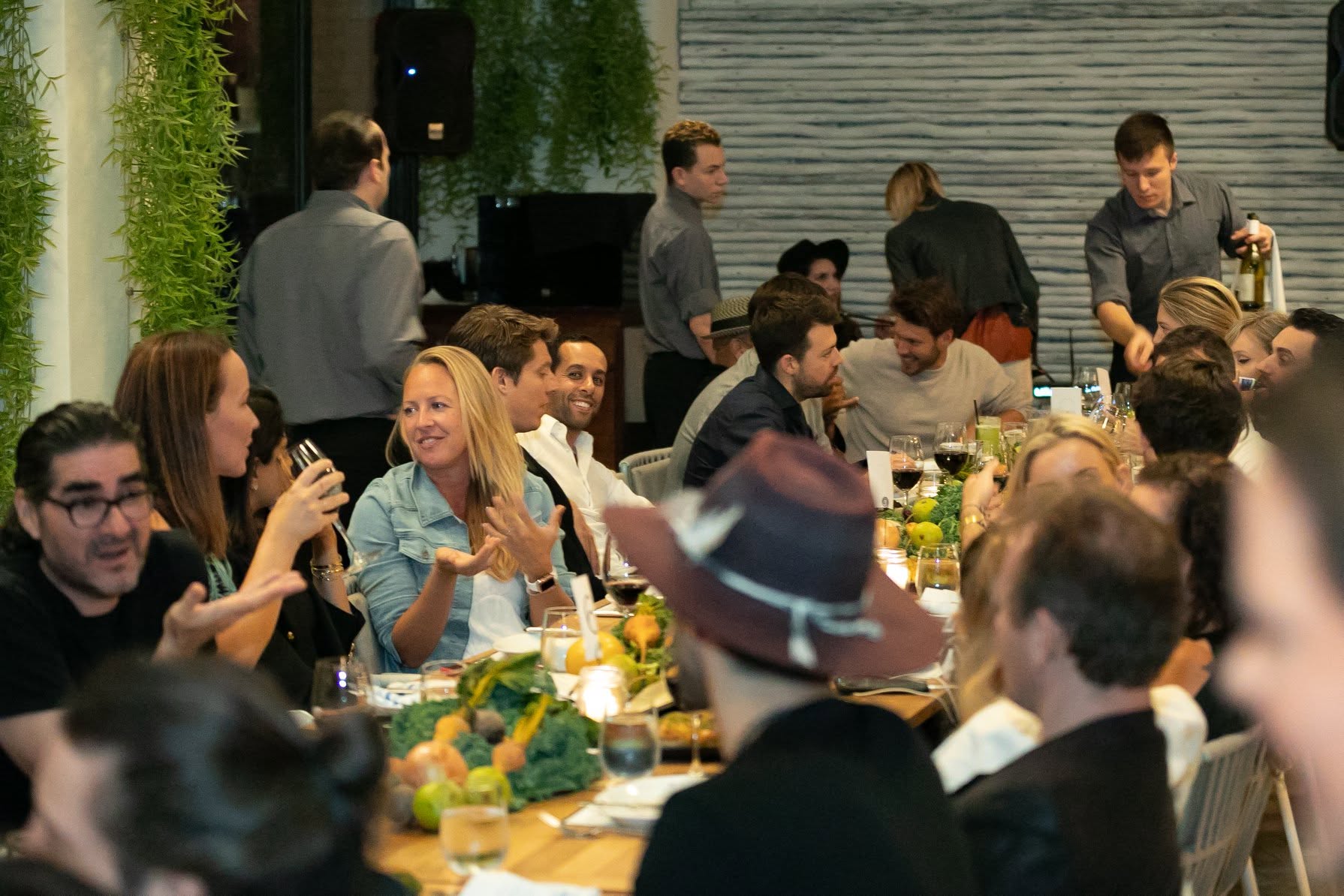 People sitting at a decorated table during a social event, some holding drinks, with a green wall and gray background.